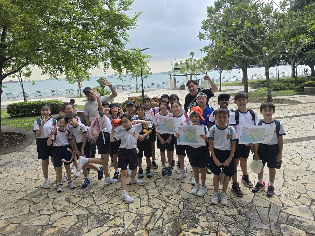 A group of school children in white uniforms holding maps pose cheerfully outdoors near a waterfront park with trees.