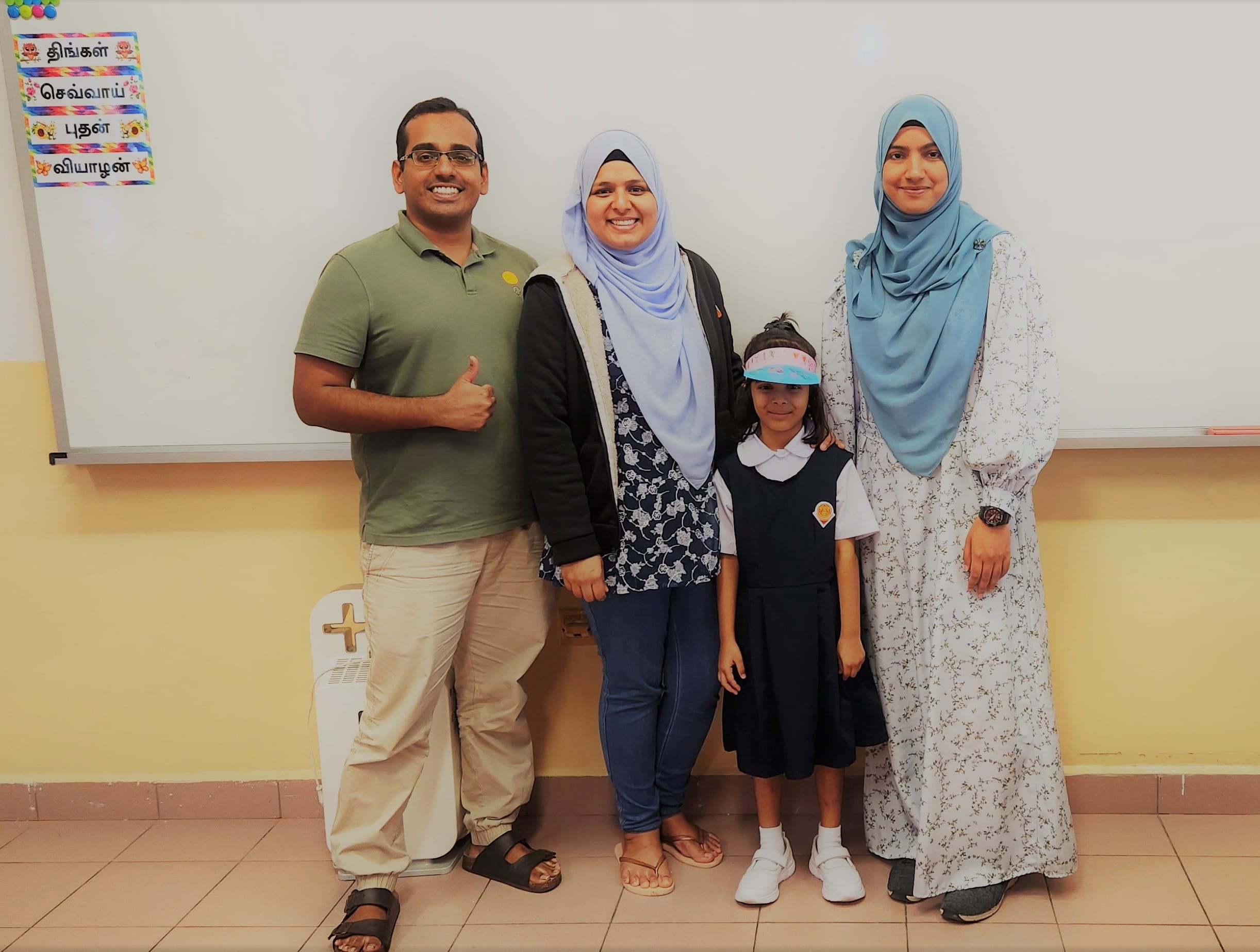 Three adults and a young girl in school uniform pose smiling in front of a whiteboard with Tamil text.
