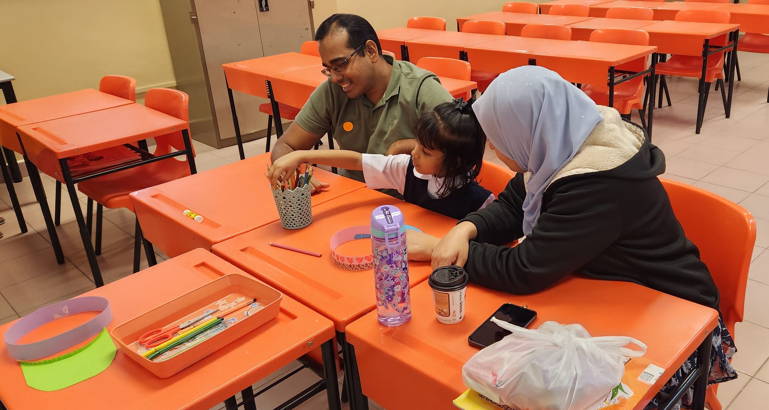 A man, young girl, and woman in a hijab sit together at orange desks in a classroom with school supplies.