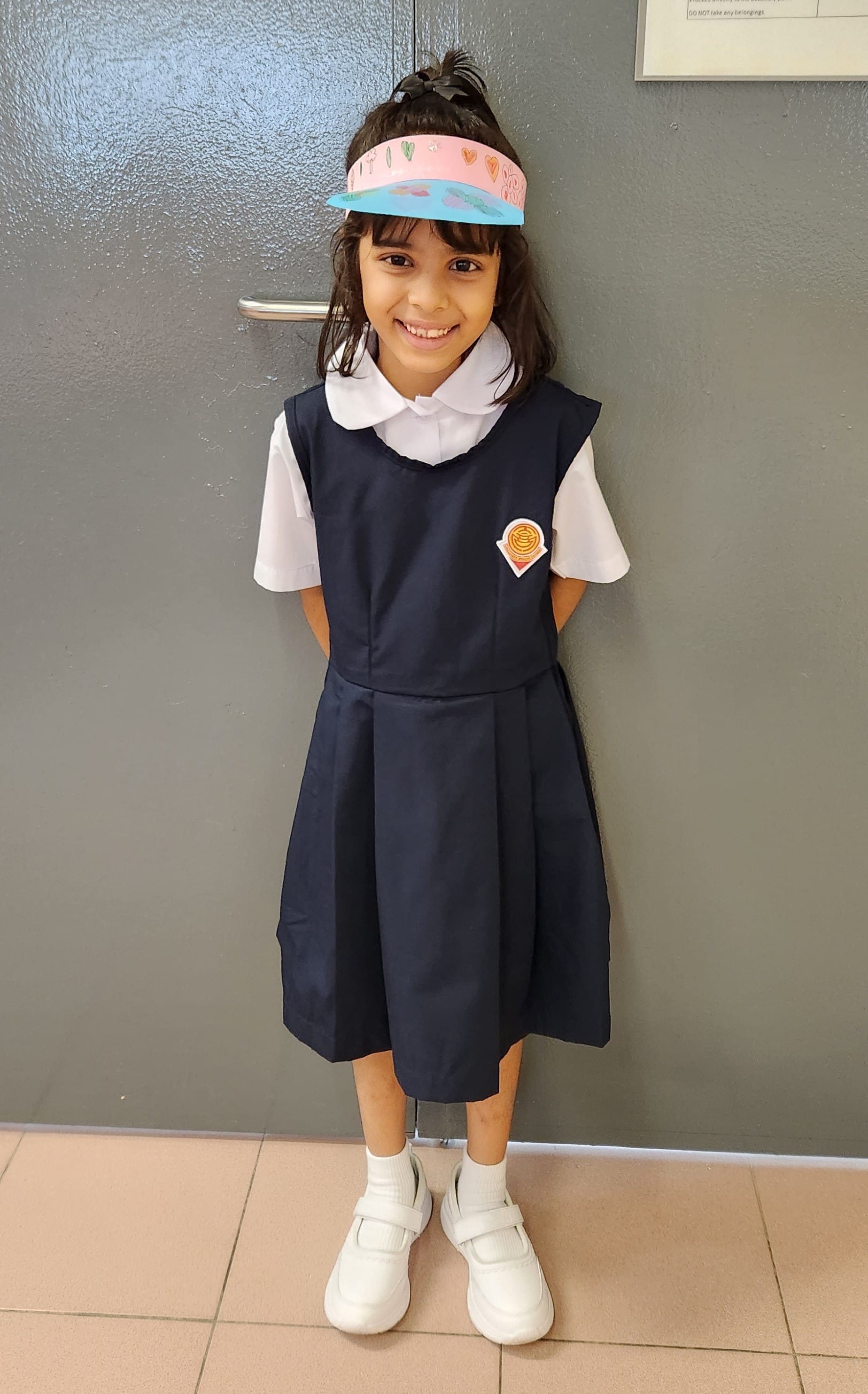Young girl in a navy school uniform with a badge, white shirt, white shoes, and a handmade paper visor, smiling.