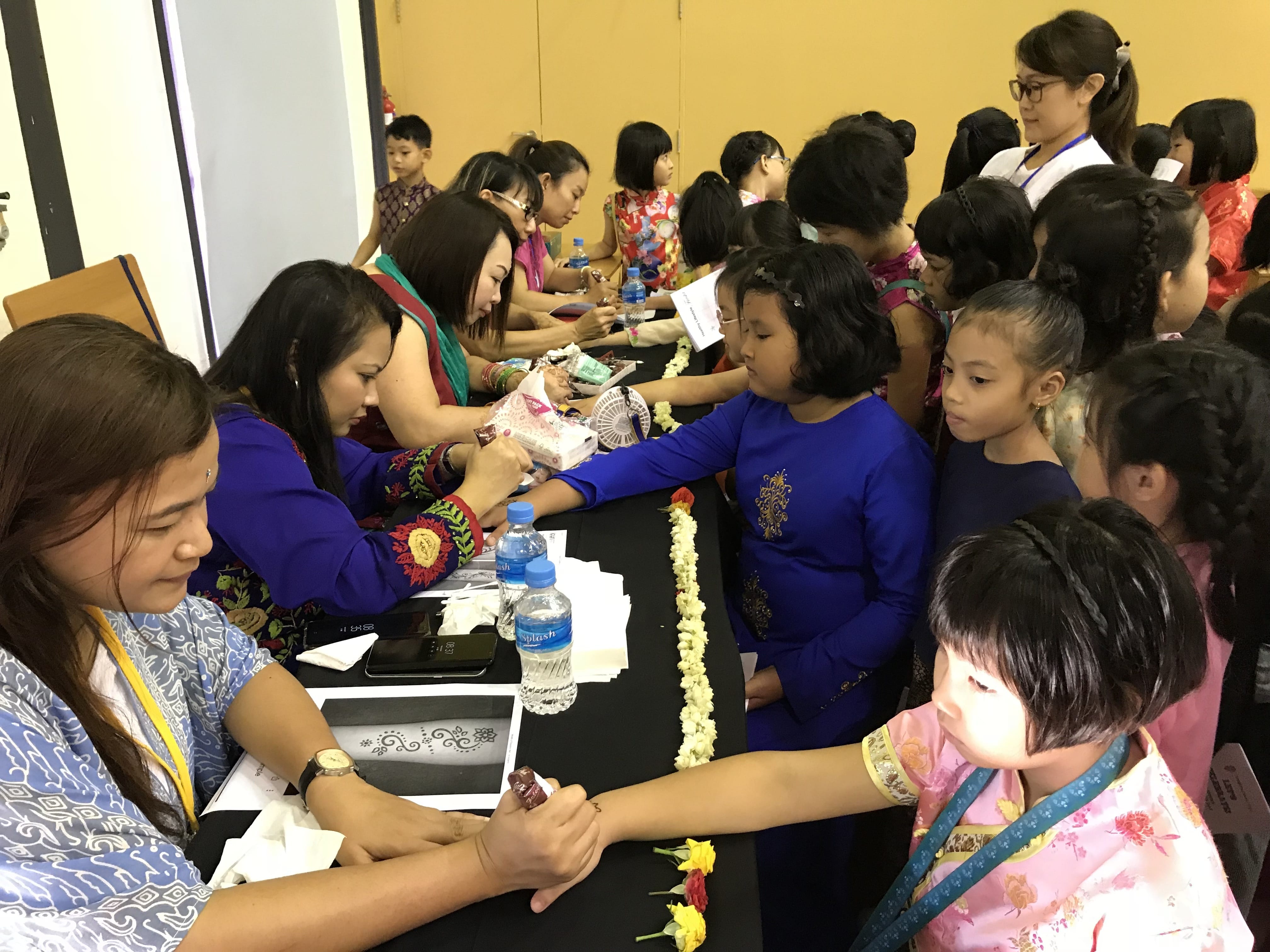 Adults apply henna to children's hands at a table decorated with flower garlands during a cultural event.