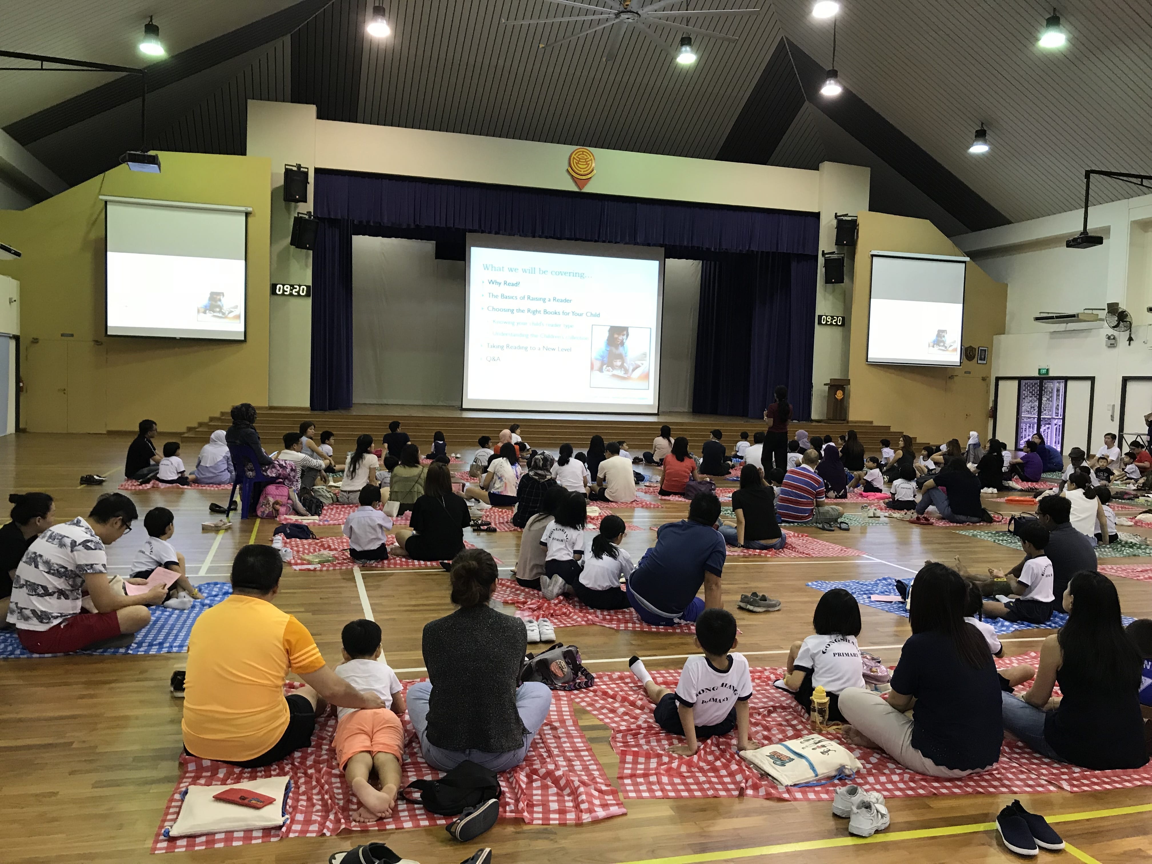 Families sit on checkered mats on a school hall floor watching a reading presentation on projected screens.