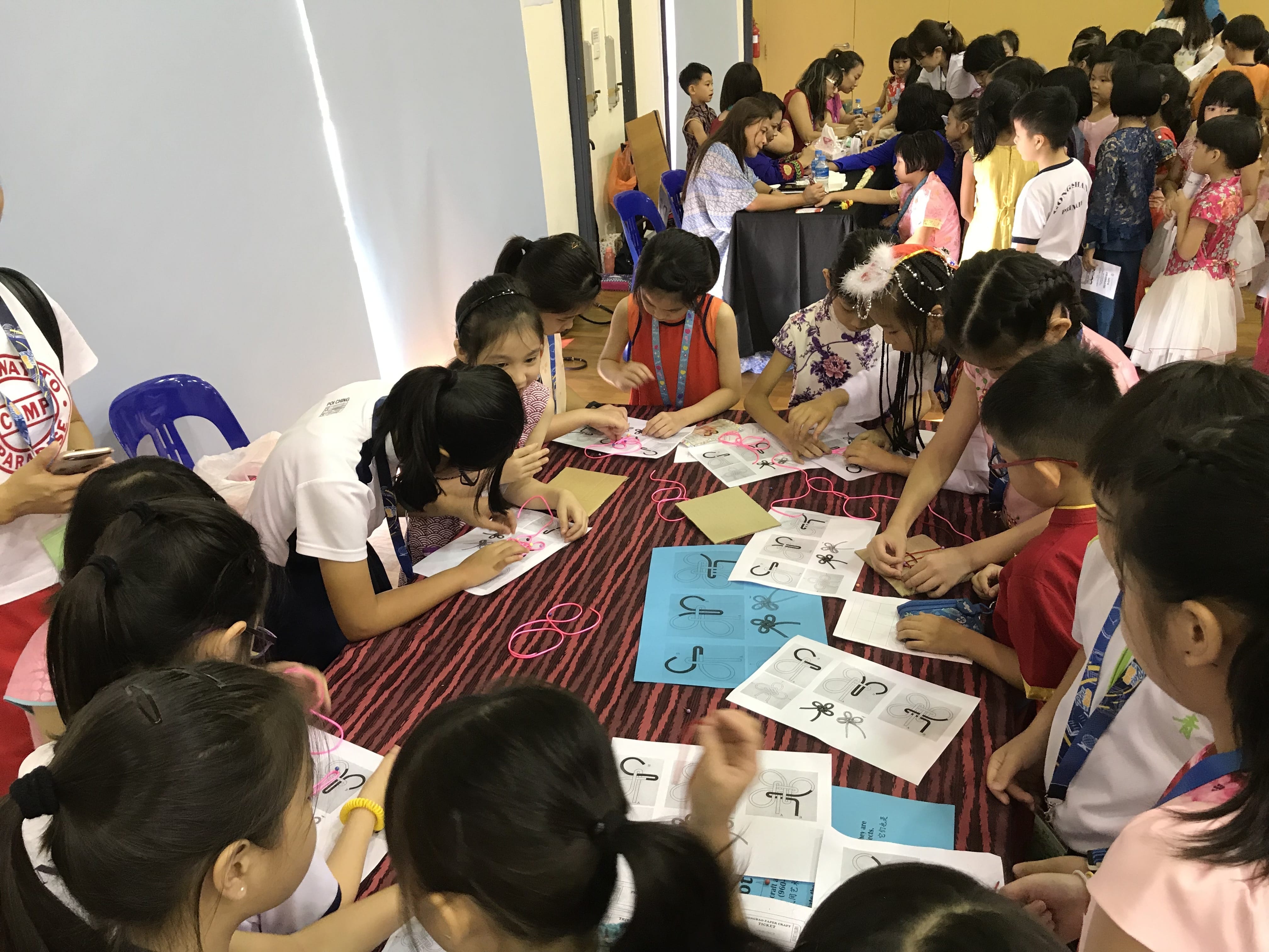 Children gather around a table doing a craft activity with printed sheets and colorful strings in a busy hall.