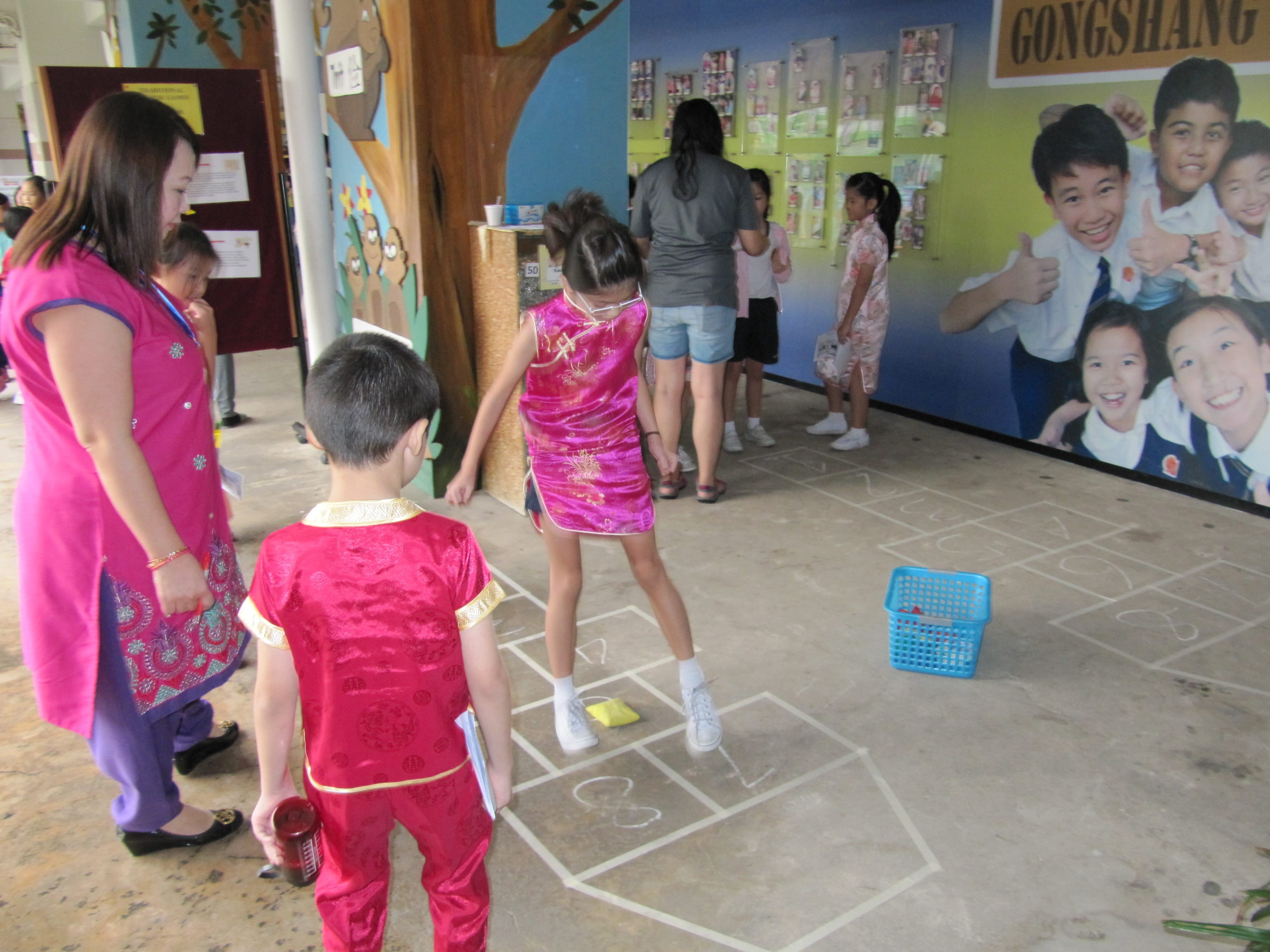 Children in traditional Chinese outfits play hopscotch on a school floor while an adult in pink watches nearby.