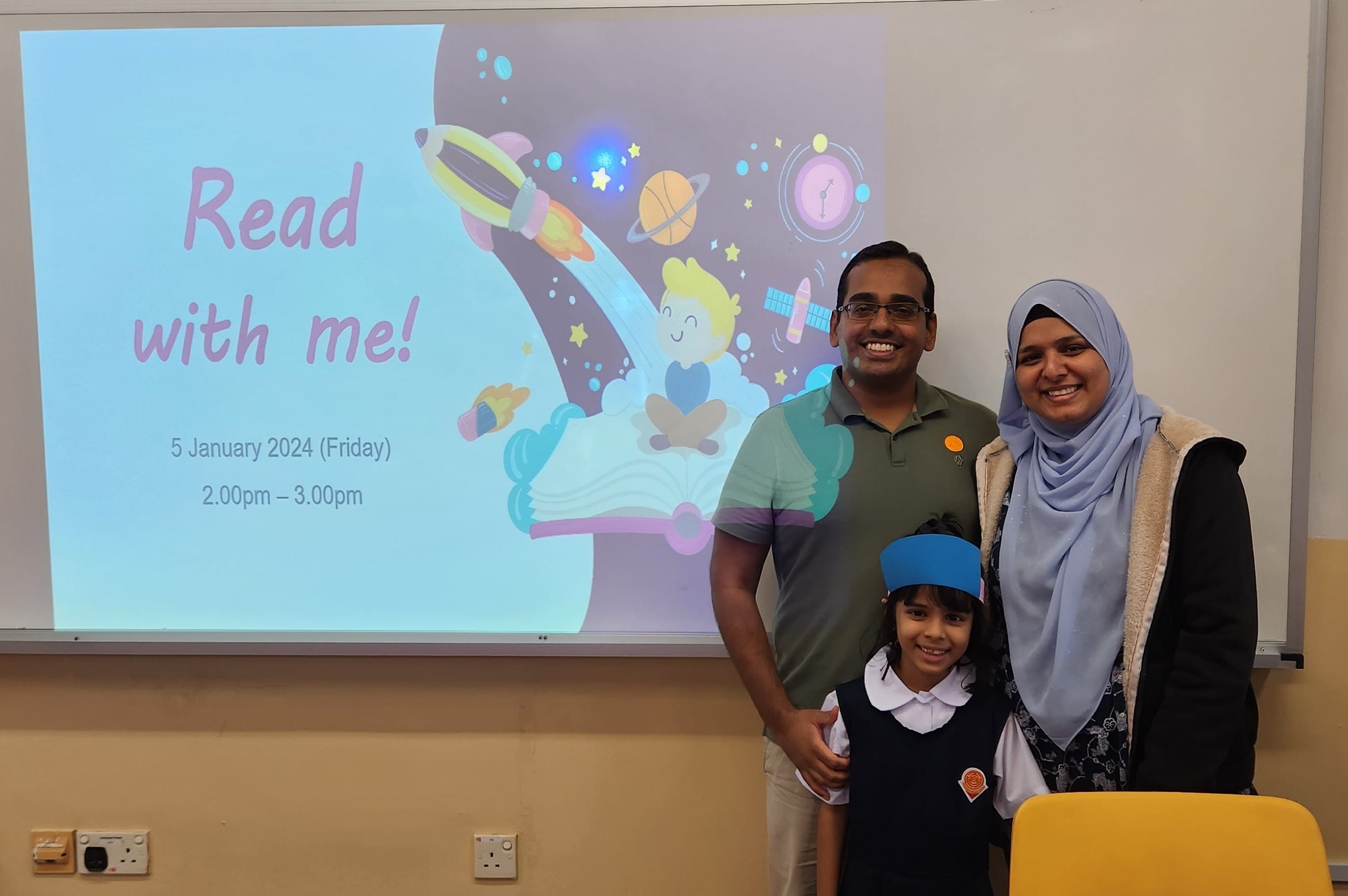 Two adults and a young girl in school uniform smile in front of a projected slide reading "Read with me! 5 January 2024."