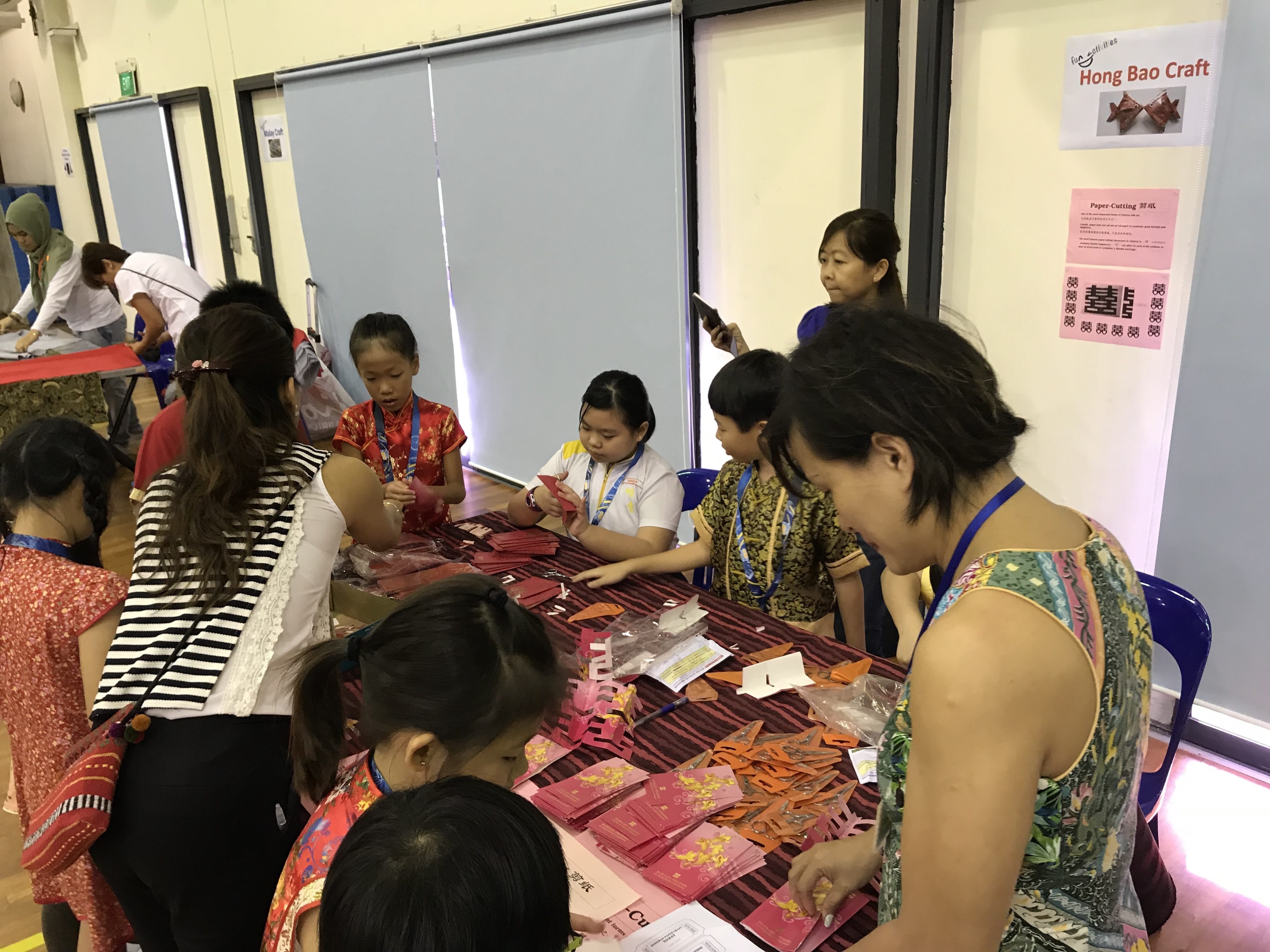 Children and adults participate in a Hong Bao Craft activity, folding red and orange paper at a table indoors.