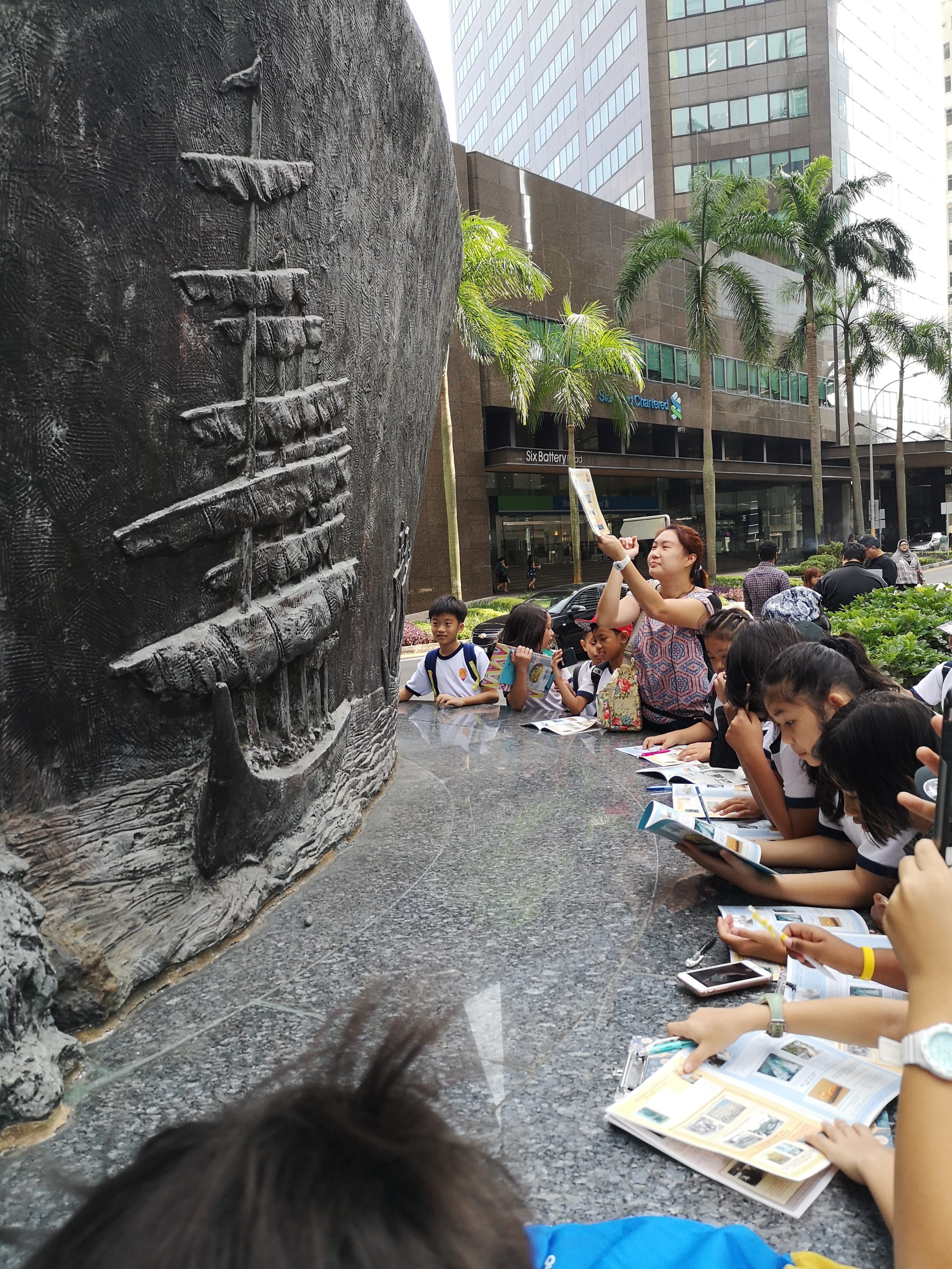 Group of students with books sit by a large ship relief sculpture while a woman takes a photo, city behind.