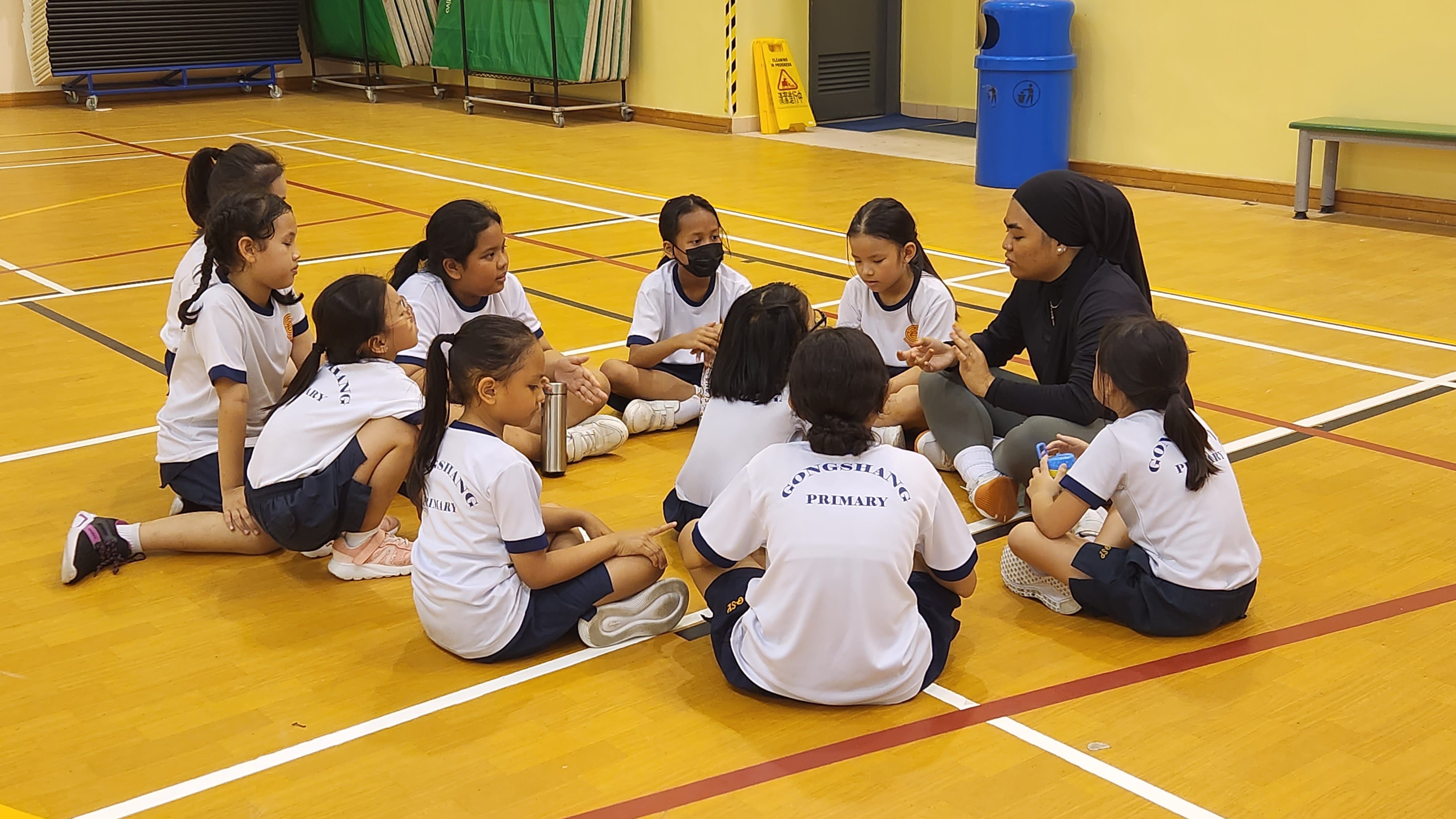 A teacher in black sits with young Gongshang Primary students in a circle on an indoor sports hall floor.