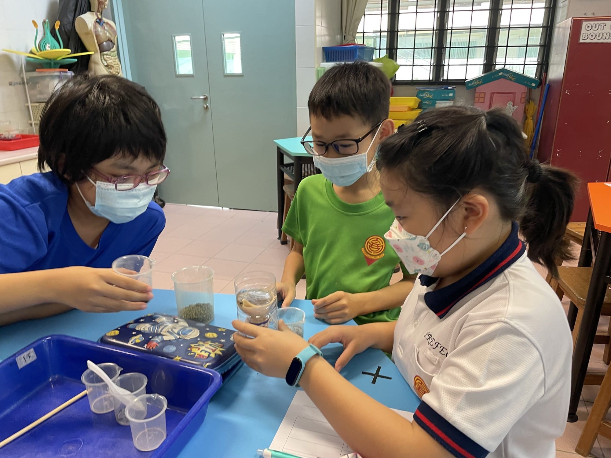 Three masked children examine cups with substances during a classroom science activity.