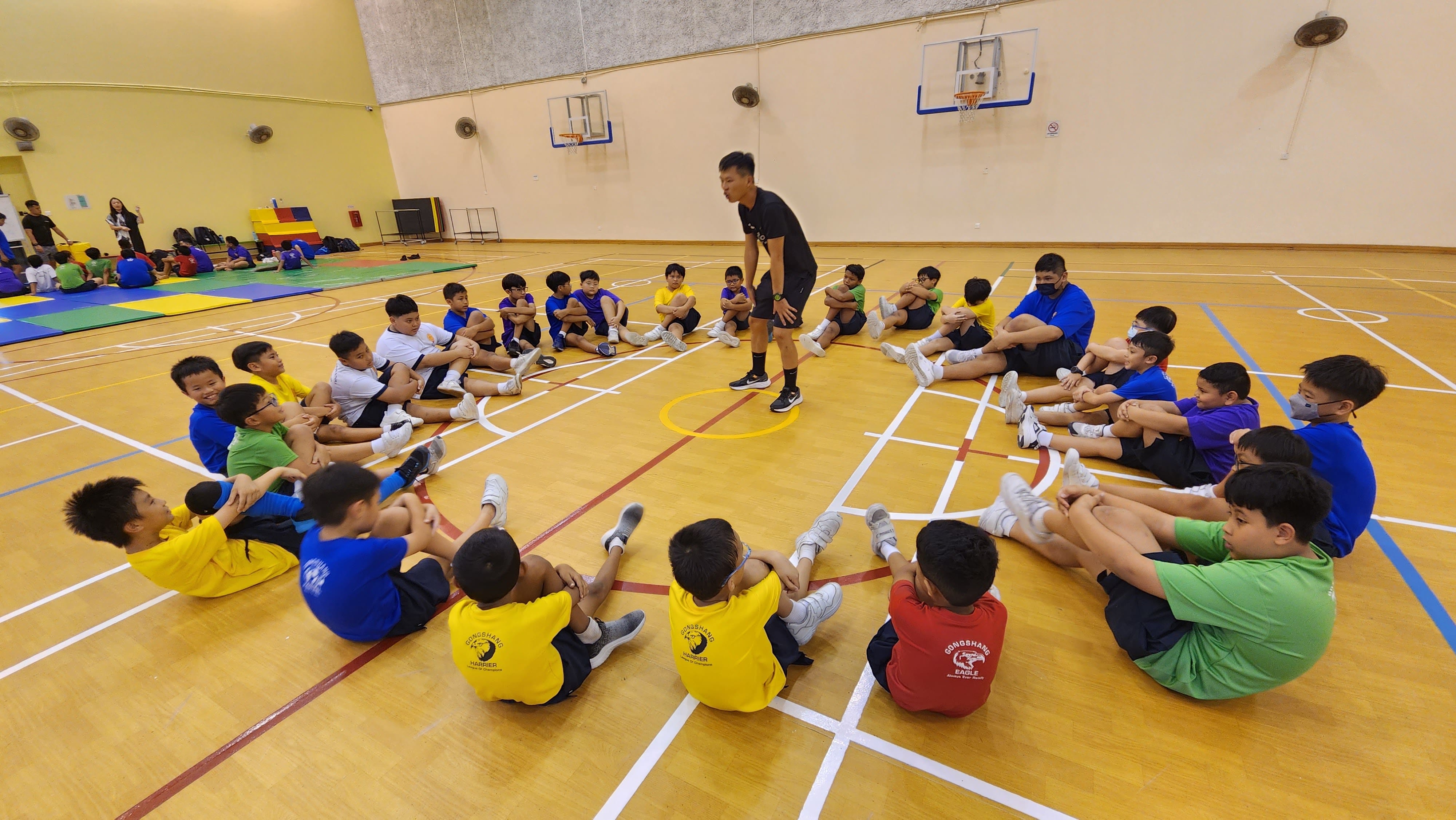 A coach in black stands before a large circle of young students seated on an indoor gymnasium floor.