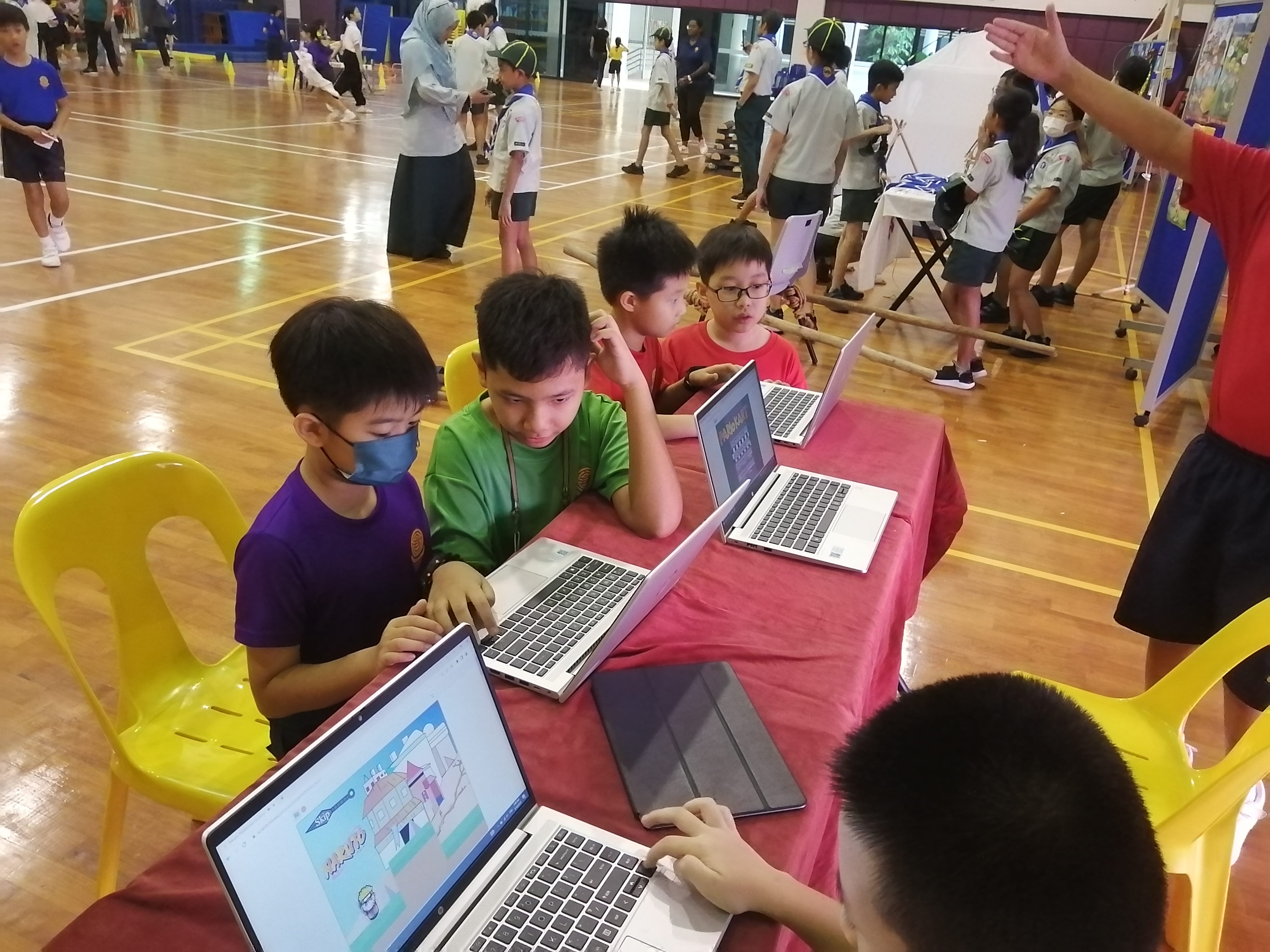 Four boys sit at a table using laptops in a gym, with other students and adults in the background.