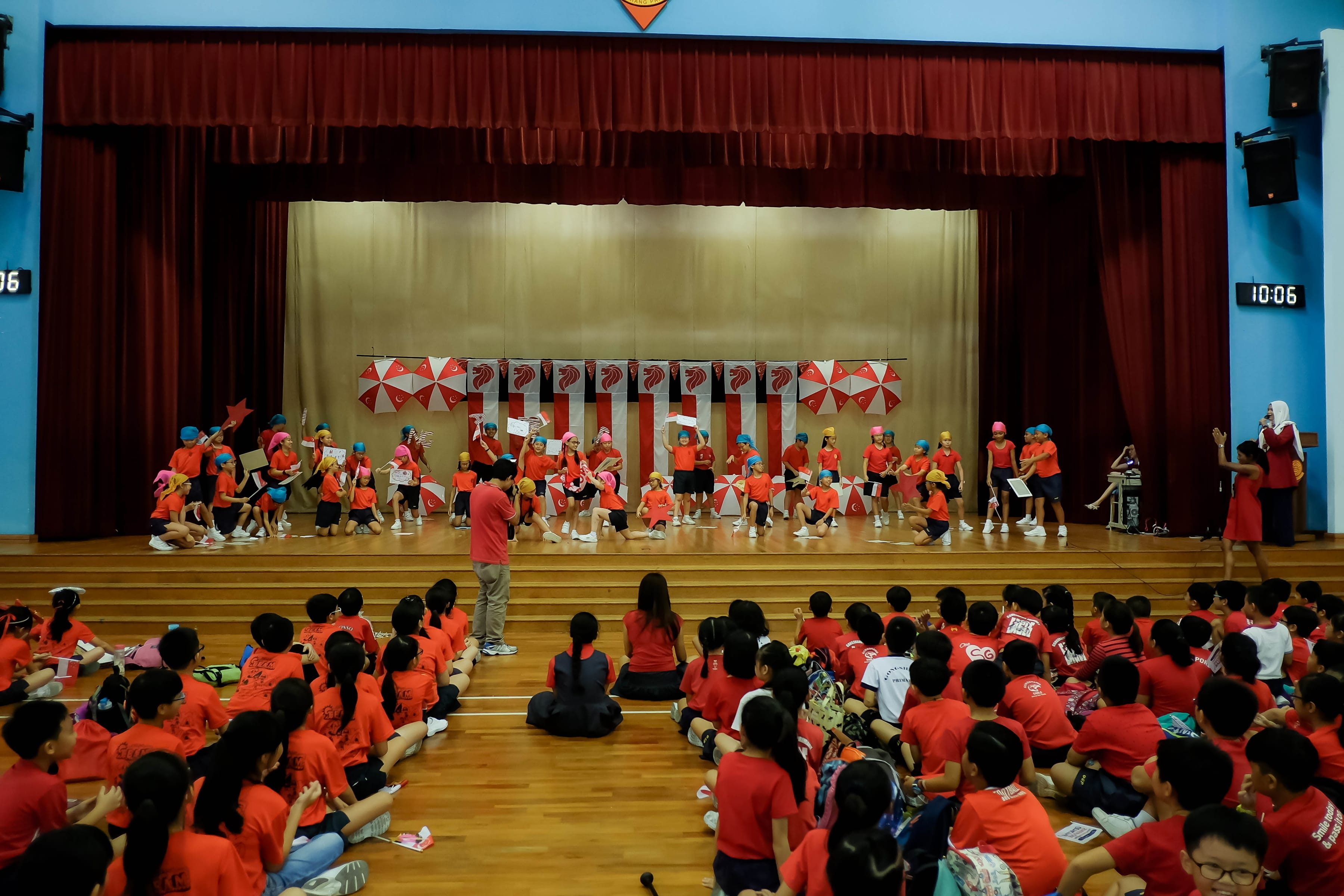 Children in red shirts perform on a stage with umbrellas while an audience of students sits on the gym floor