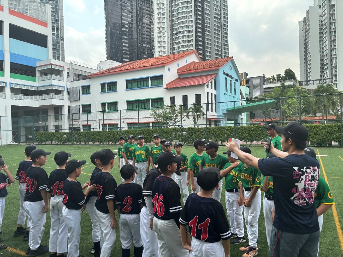 Two youth baseball teams in black and green uniforms gather on a field near a school building with tall towers behind.