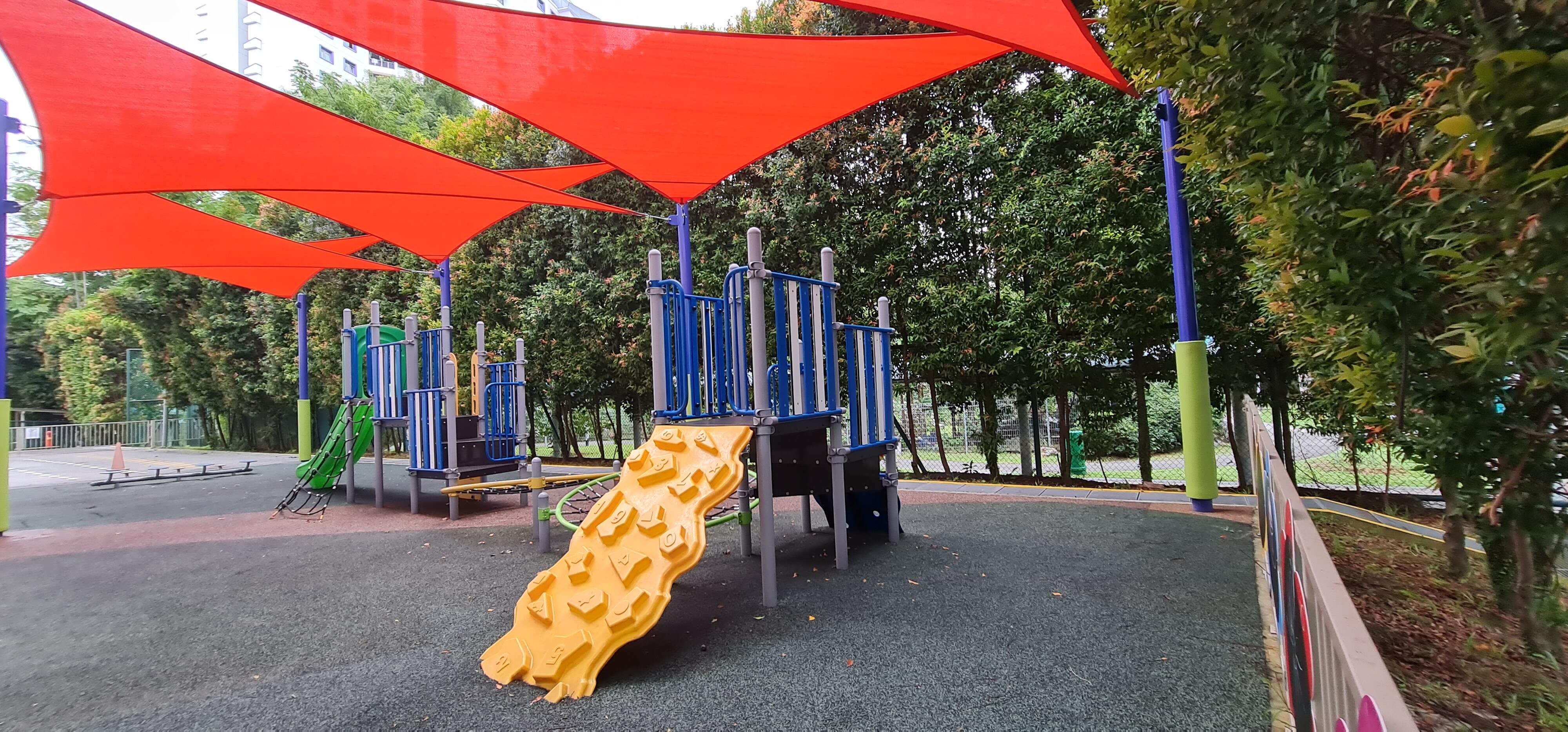 Outdoor playground with a yellow textured slide, blue climbing frame, and large red triangular shade sails overhead.
