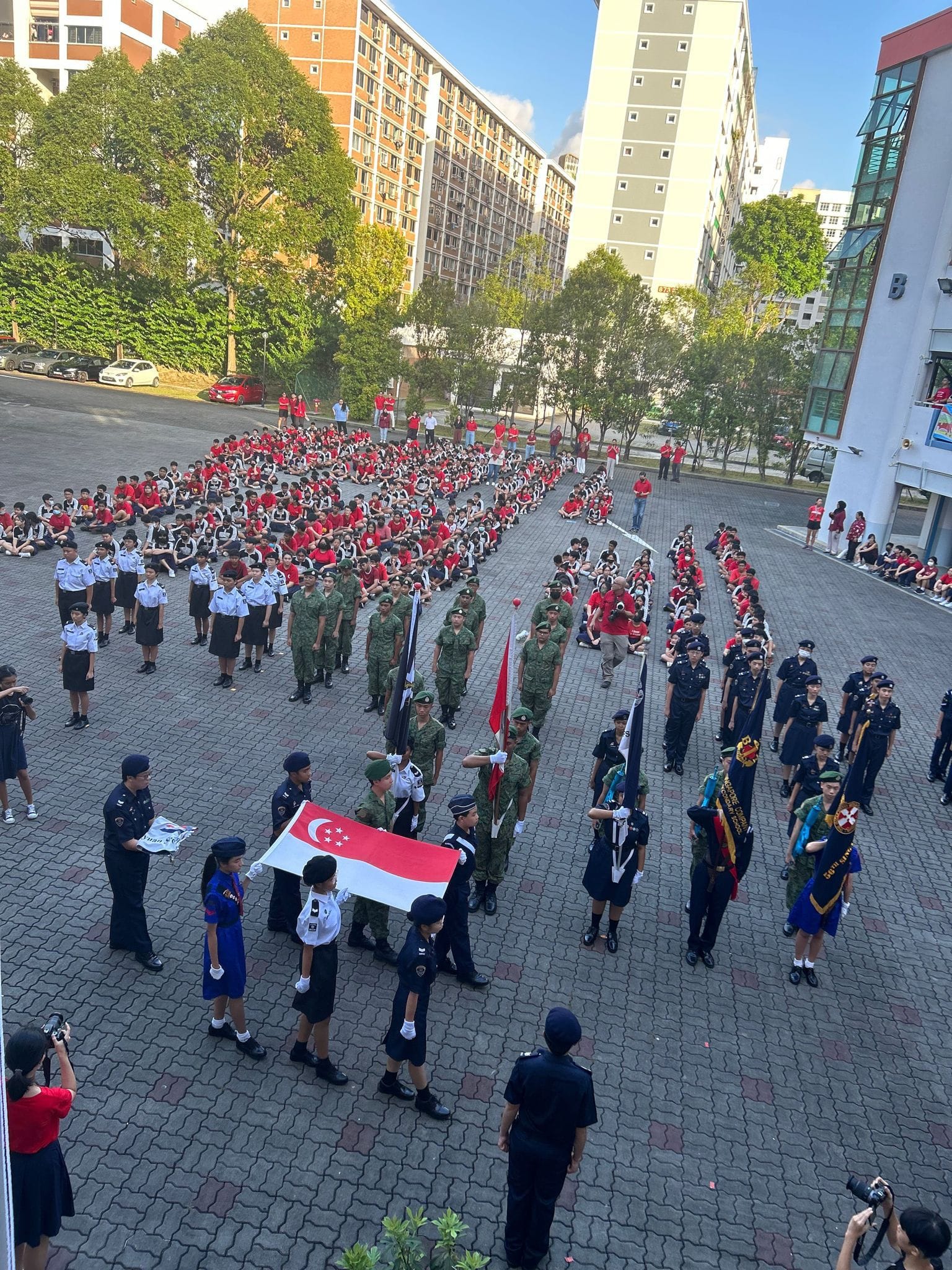 Students in uniforms stand in formation at a school parade, with the Singapore flag being carried at the front.