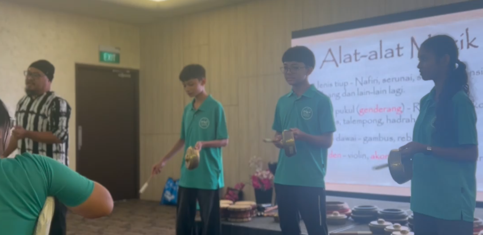 Three students in teal shirts play small percussion instruments beside a screen titled Alat-alat Muzik.