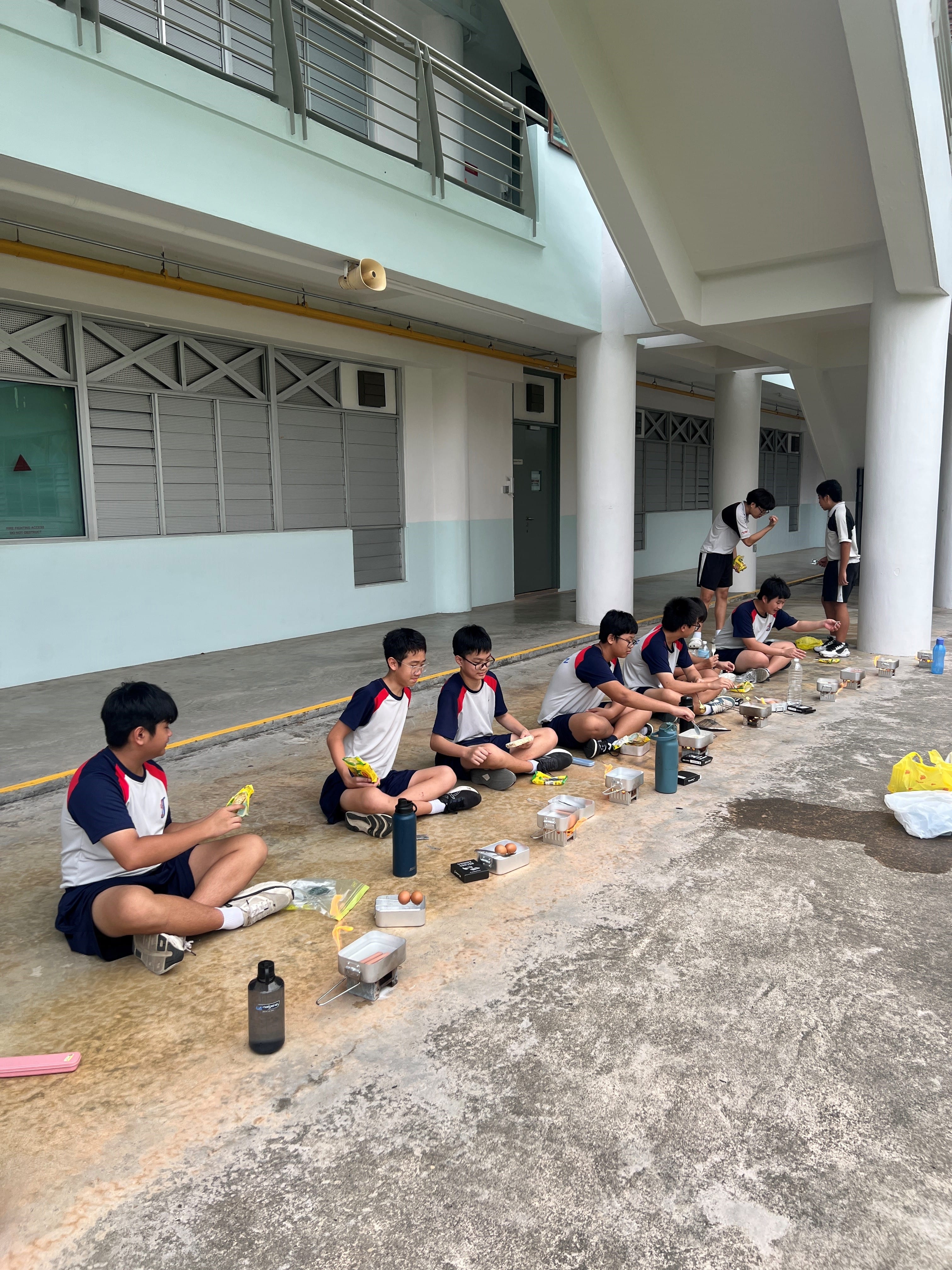 Students in school uniforms sit on a covered outdoor corridor floor with food containers and water bottles in front of them.