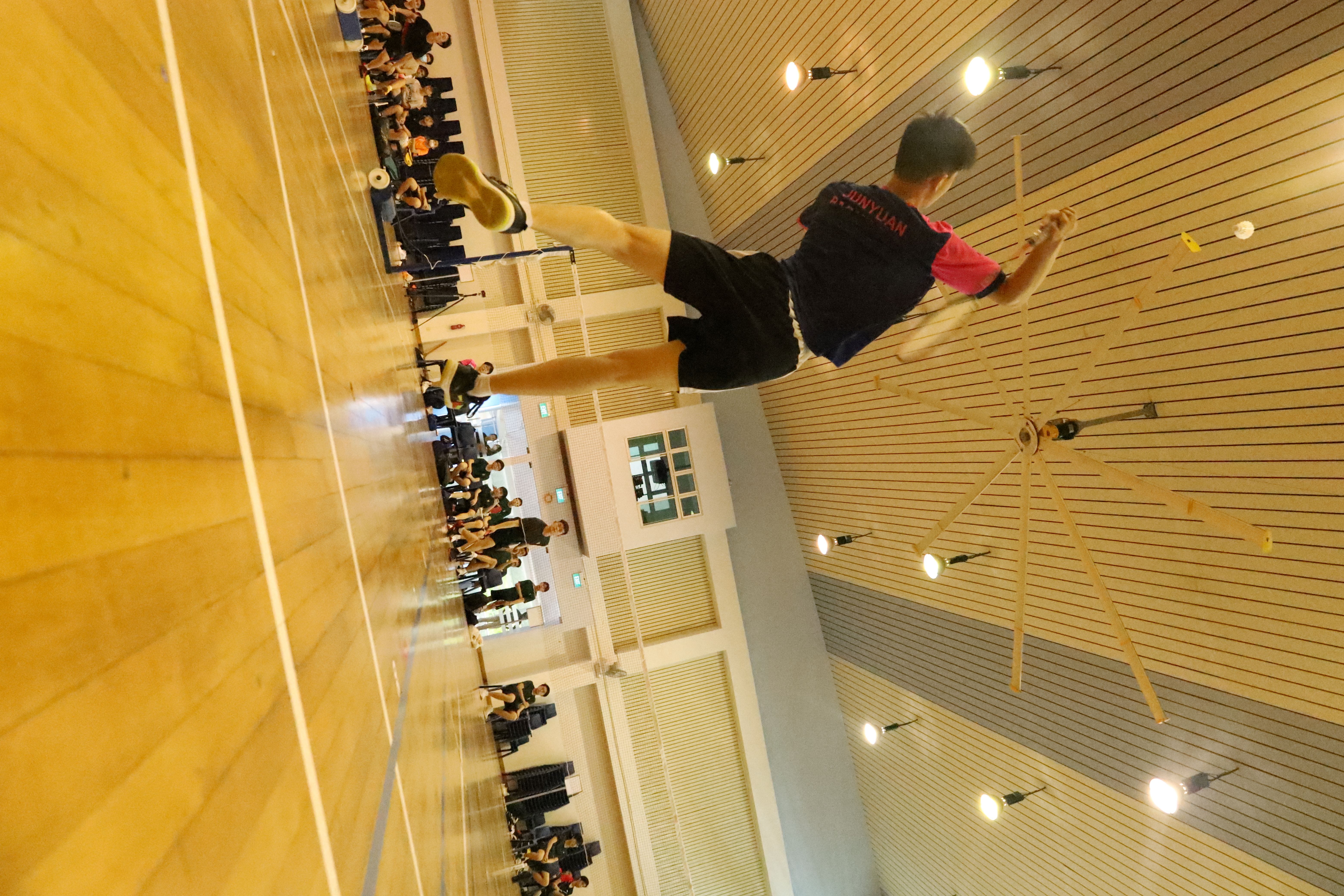 A badminton player jumps mid-air to smash, viewed from the court floor, with spectators seated along the walls.
