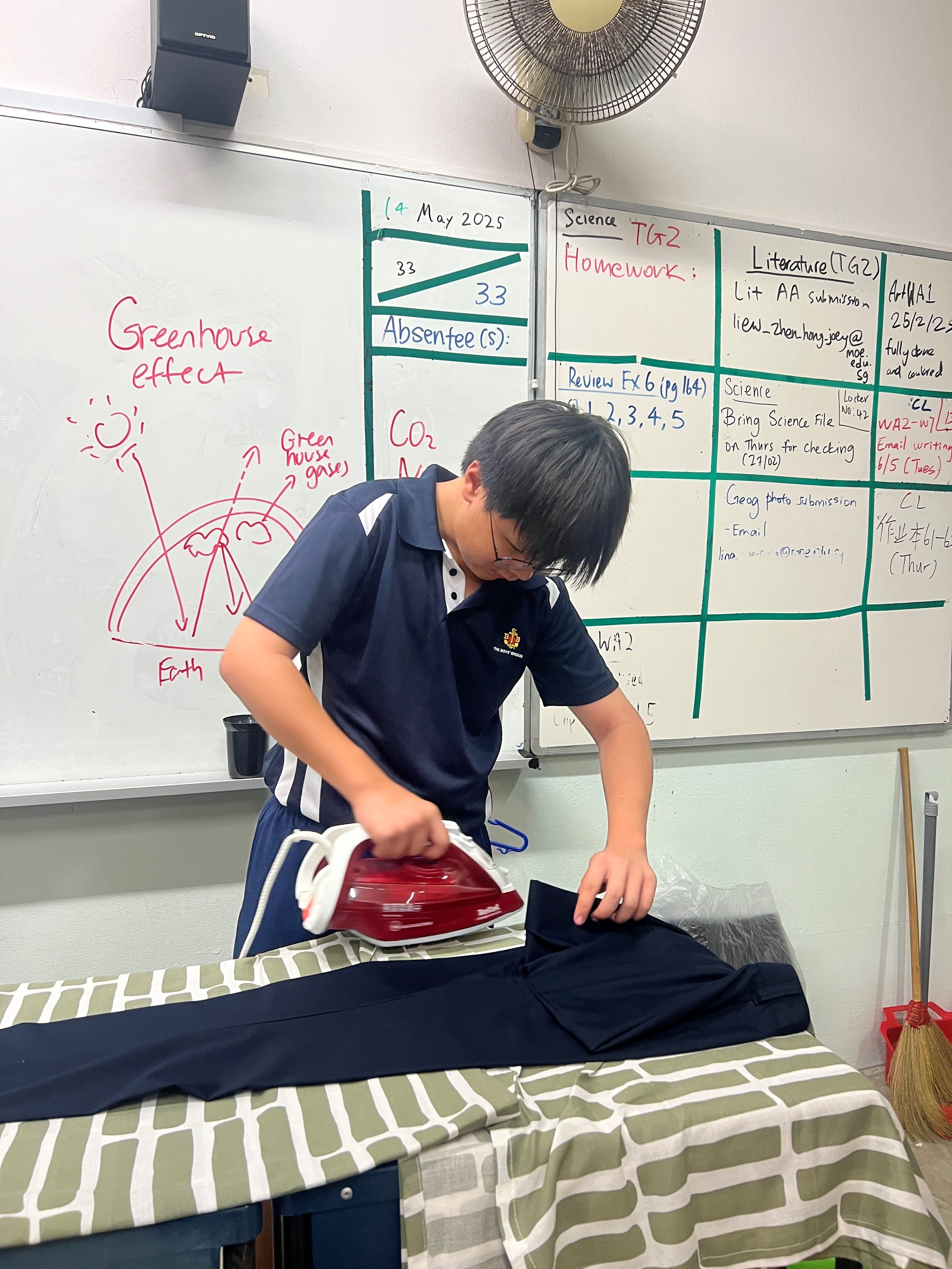 A student in a navy uniform irons dark pants on a board in a classroom with a whiteboard showing greenhouse effect notes.