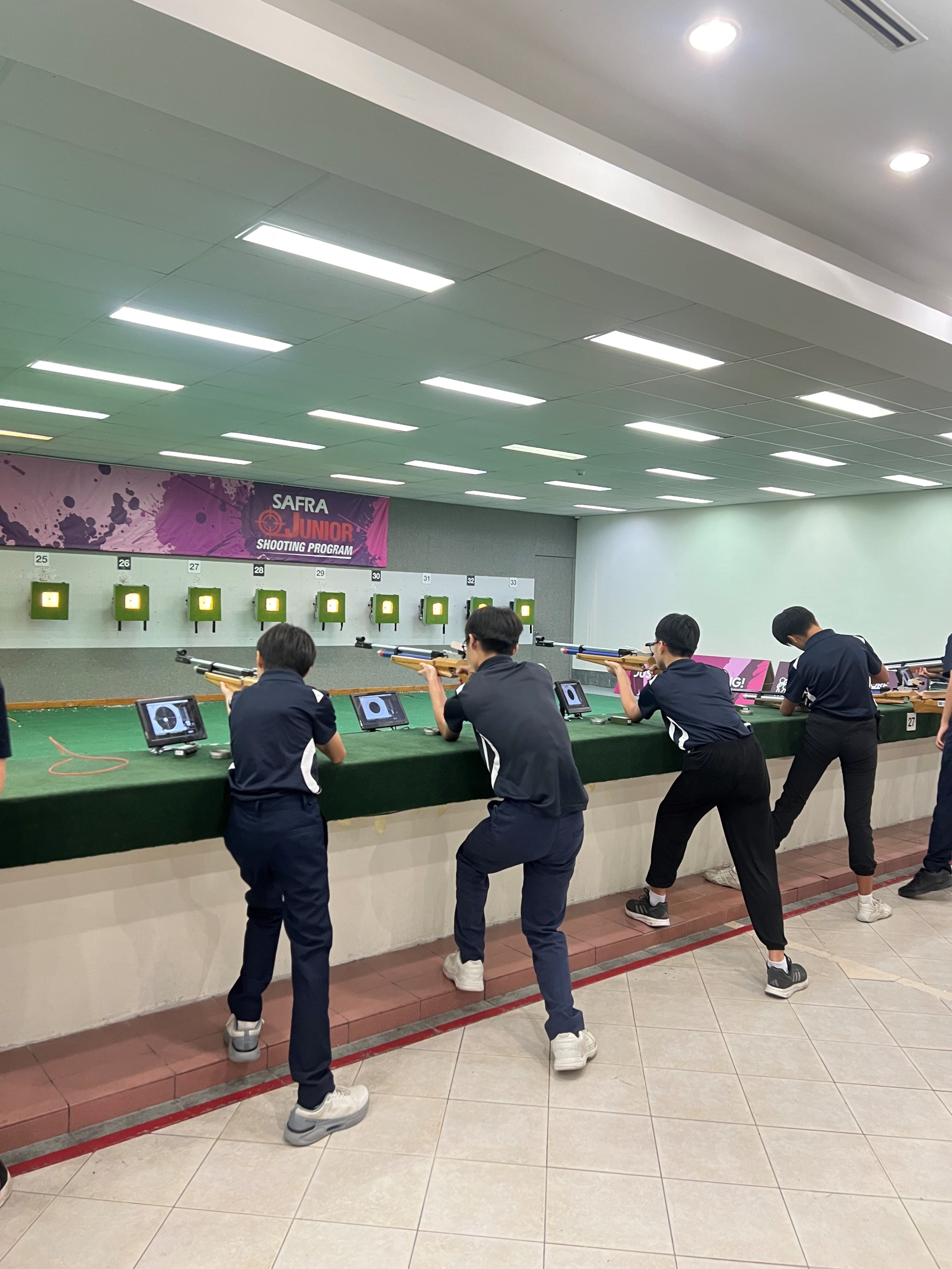 Several young shooters in black uniforms aim rifles at targets in an indoor SAFRA Junior Shooting Program range.