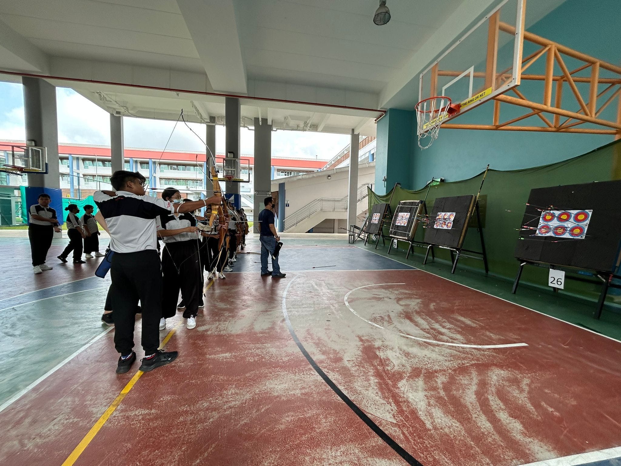 Students in black and white uniforms practice archery on an indoor basketball court, aiming at circular targets on stands.