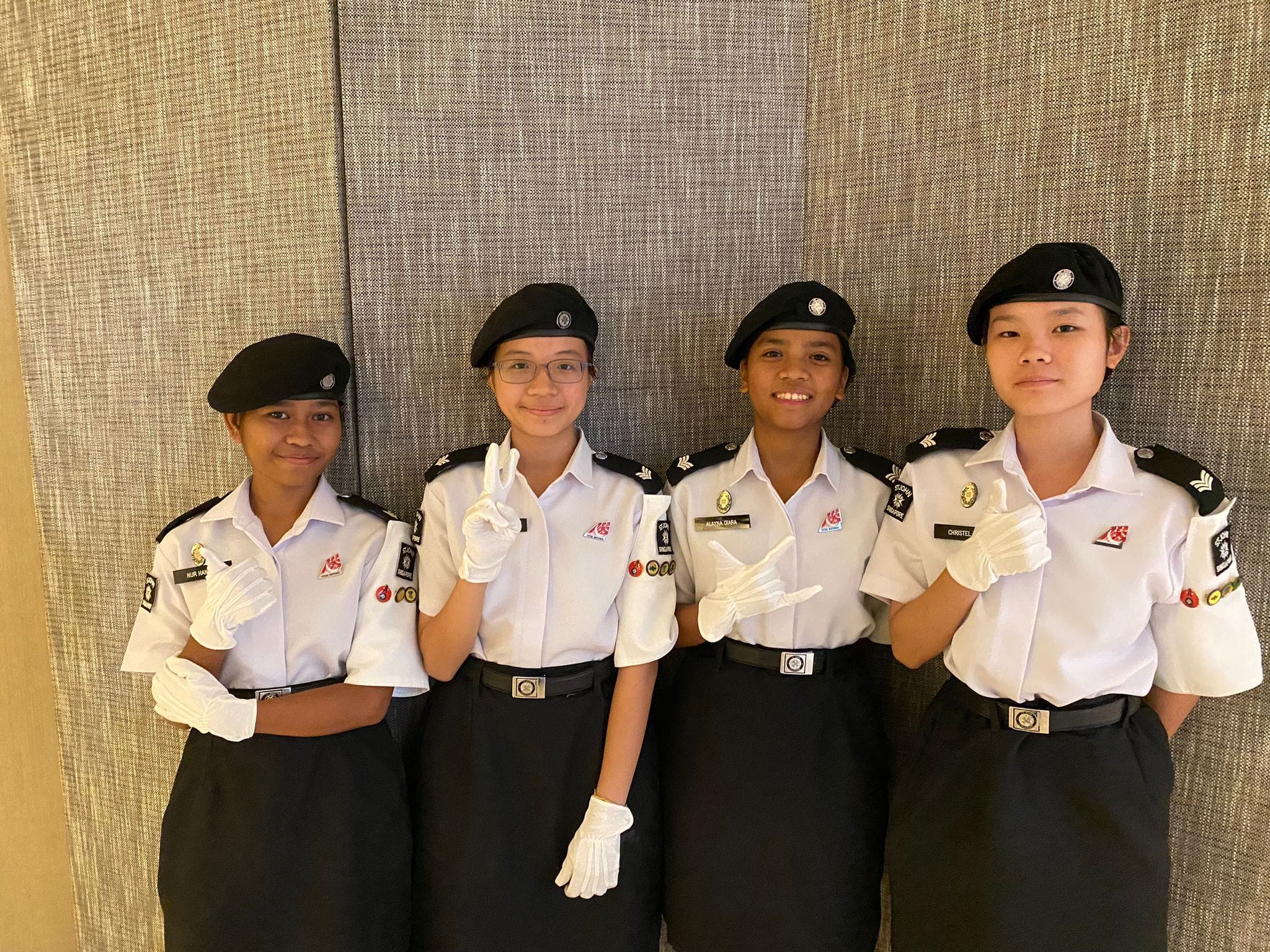 Four young women in white and black uniforms with black berets and white gloves pose smiling together.