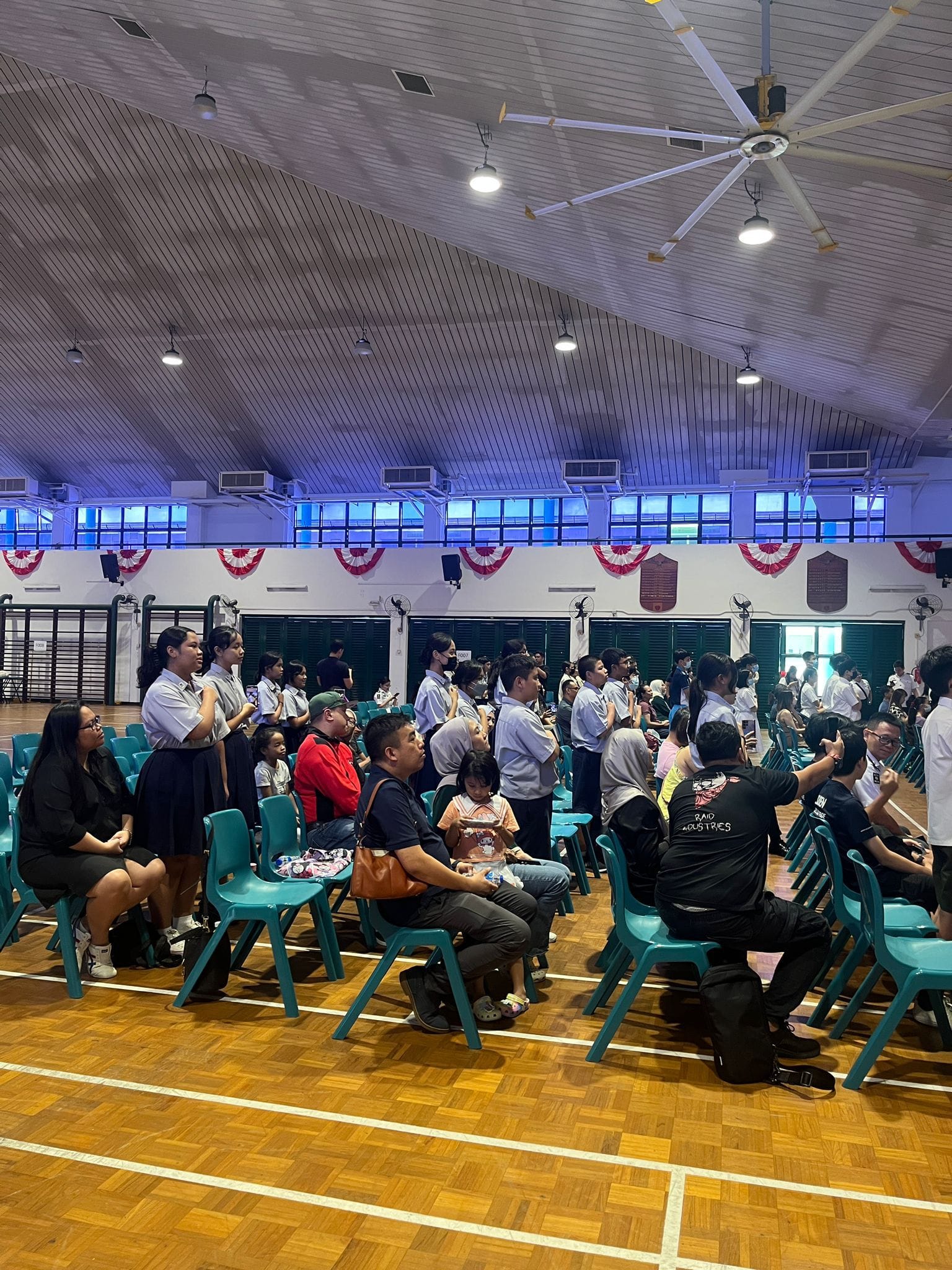 A crowd of people seated on teal chairs in a school hall decorated with red bunting and blue lights.