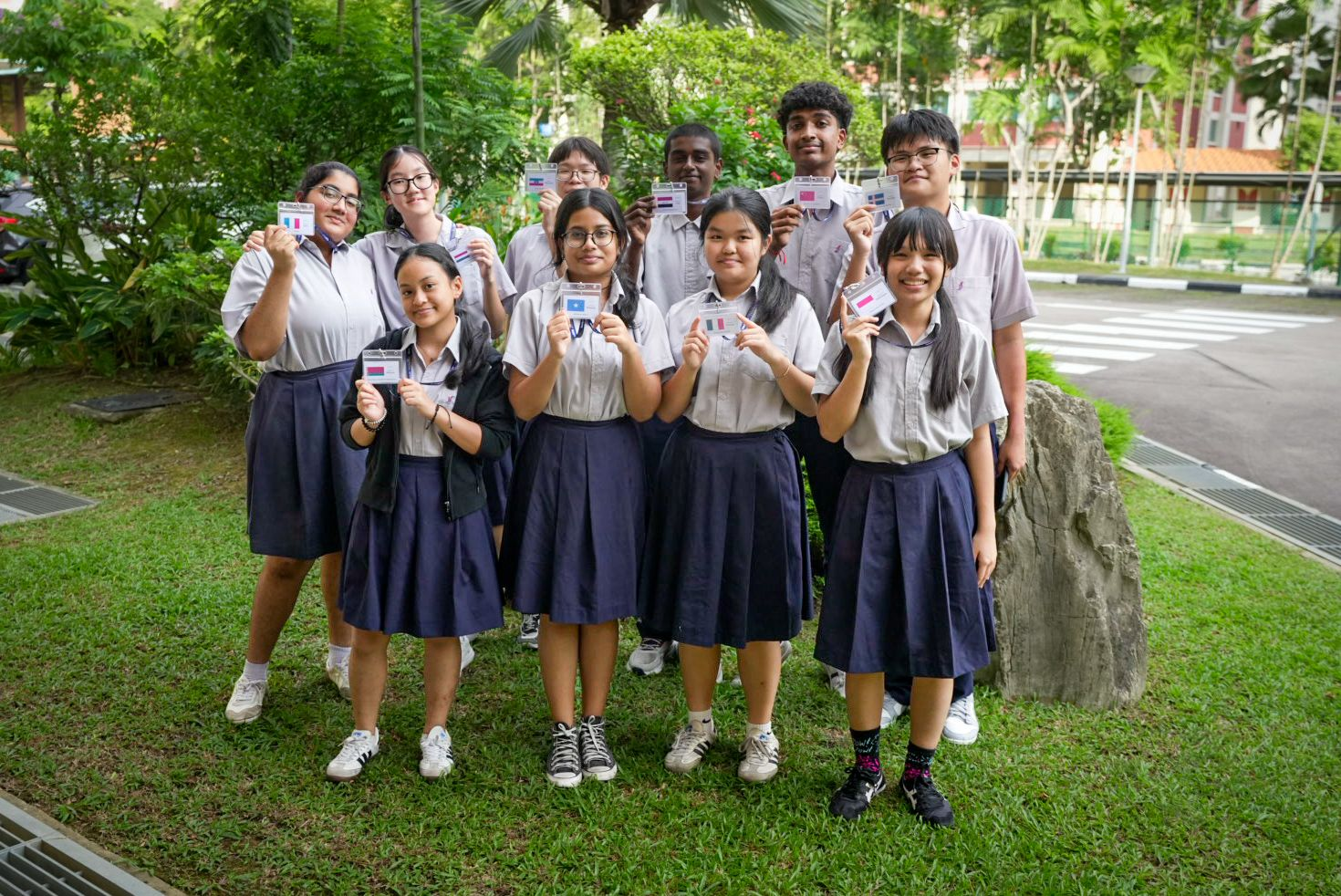 Ten students in school uniforms stand on grass outdoors, each holding up a small card with a country flag.