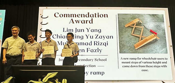 Four people stand on stage holding certificates beside a Commendation Award board with student names listed.