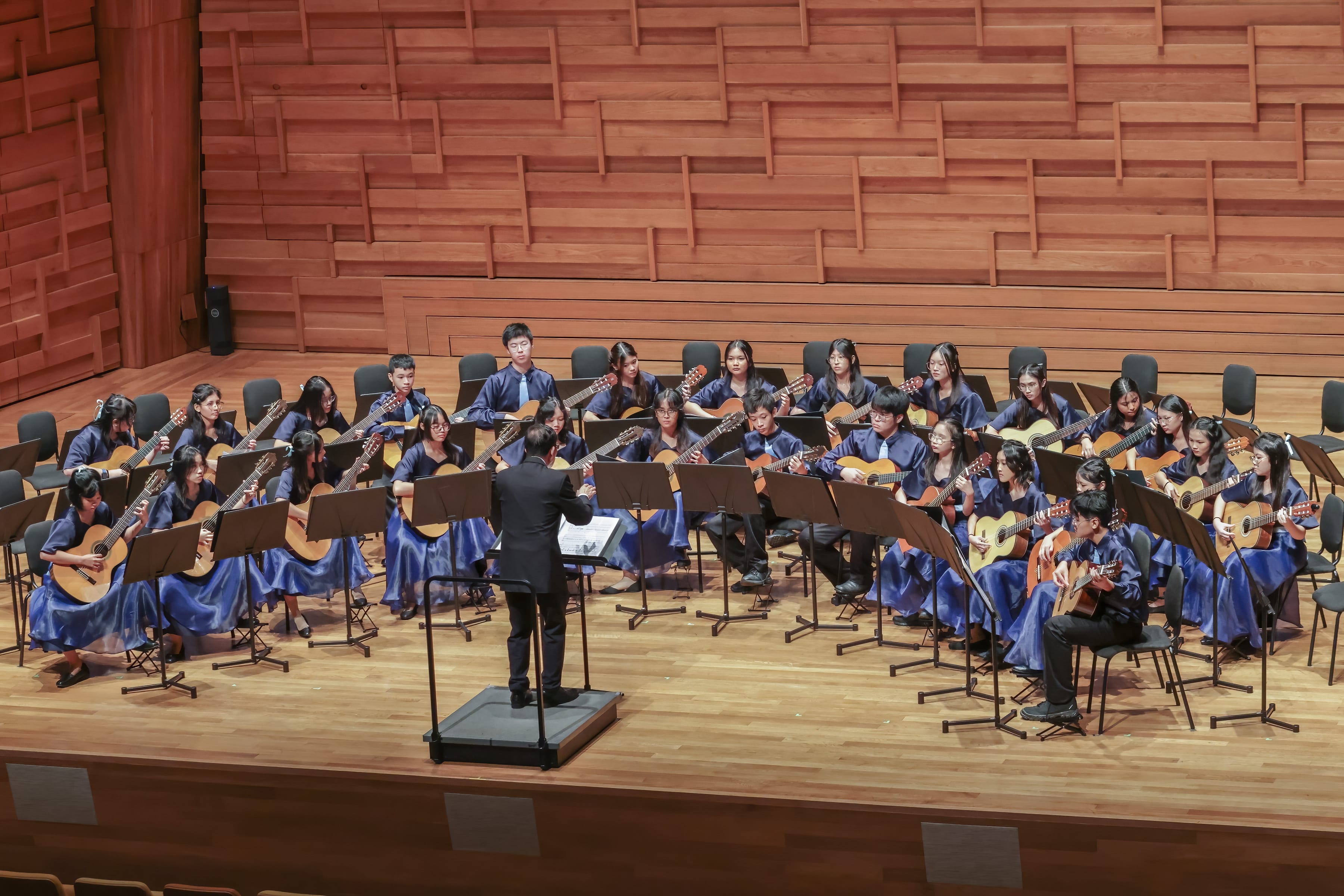A conductor leads a large youth guitar ensemble in blue robes performing on a wooden concert hall stage.