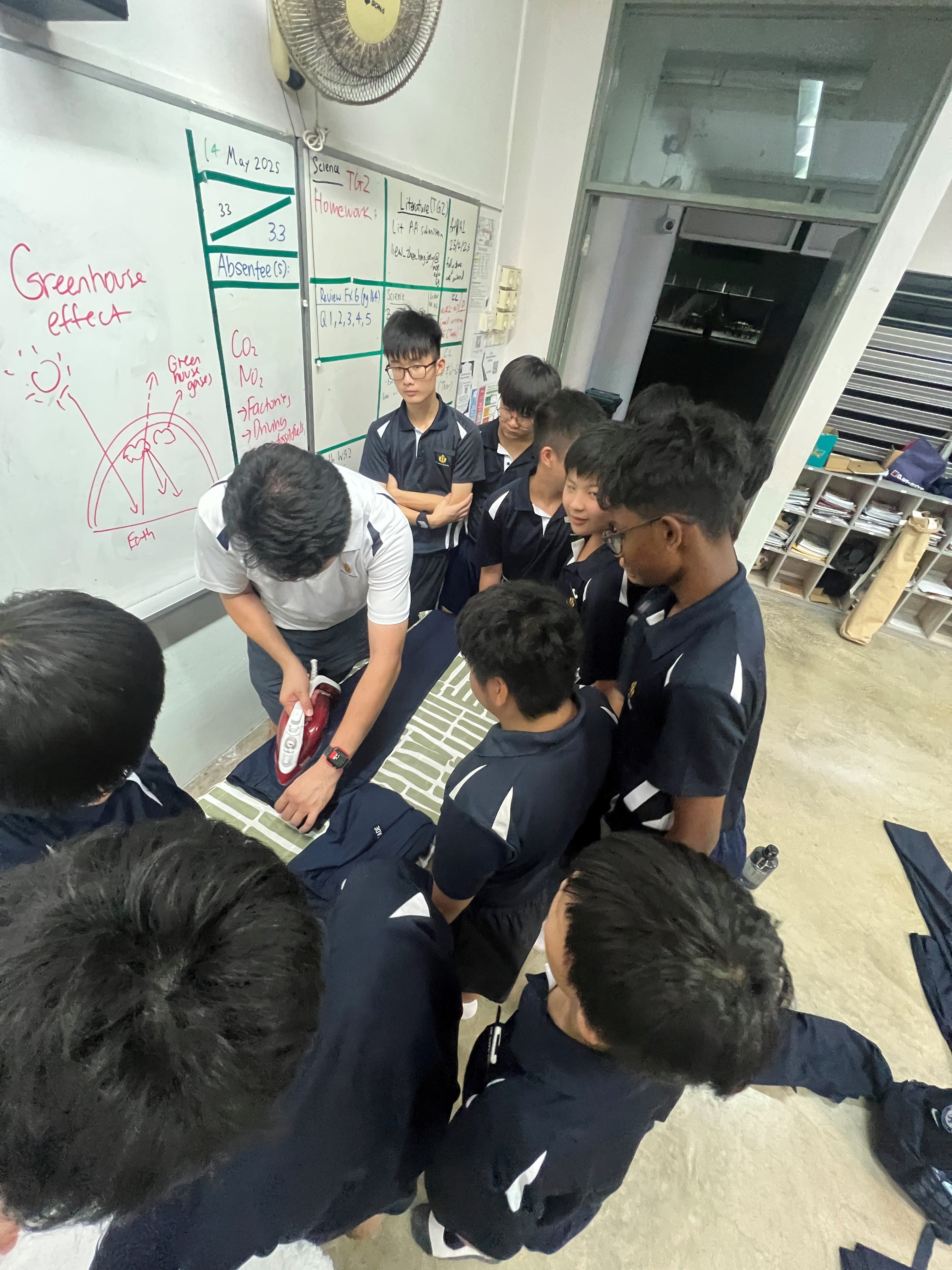 Students in navy uniforms gather around a classmate using a clothes iron on fabric near a whiteboard showing greenhouse effect notes.