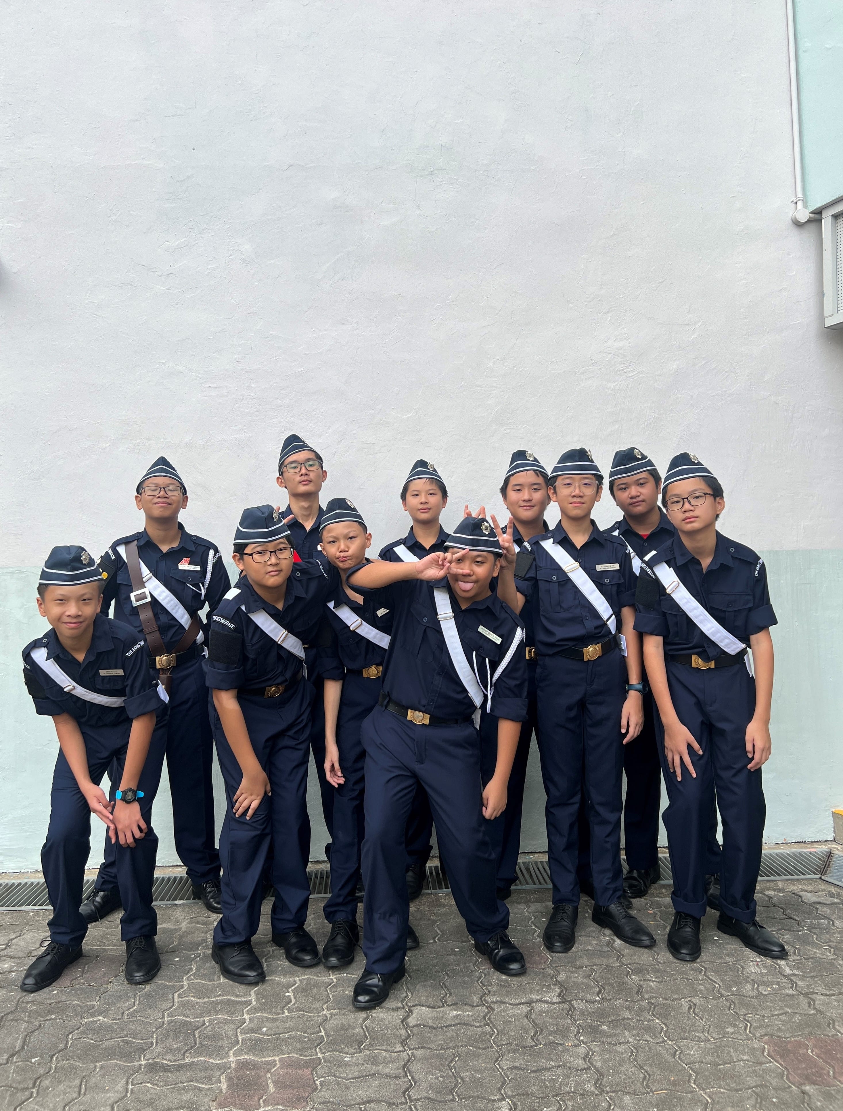 A group of young students in navy blue uniforms with white sashes and caps posing together outdoors.