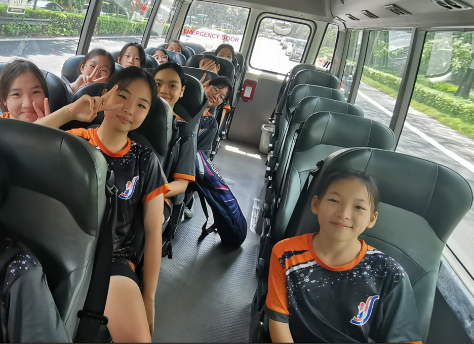 Group of young girls in matching black and orange sports jerseys smiling and posing on a moving bus.