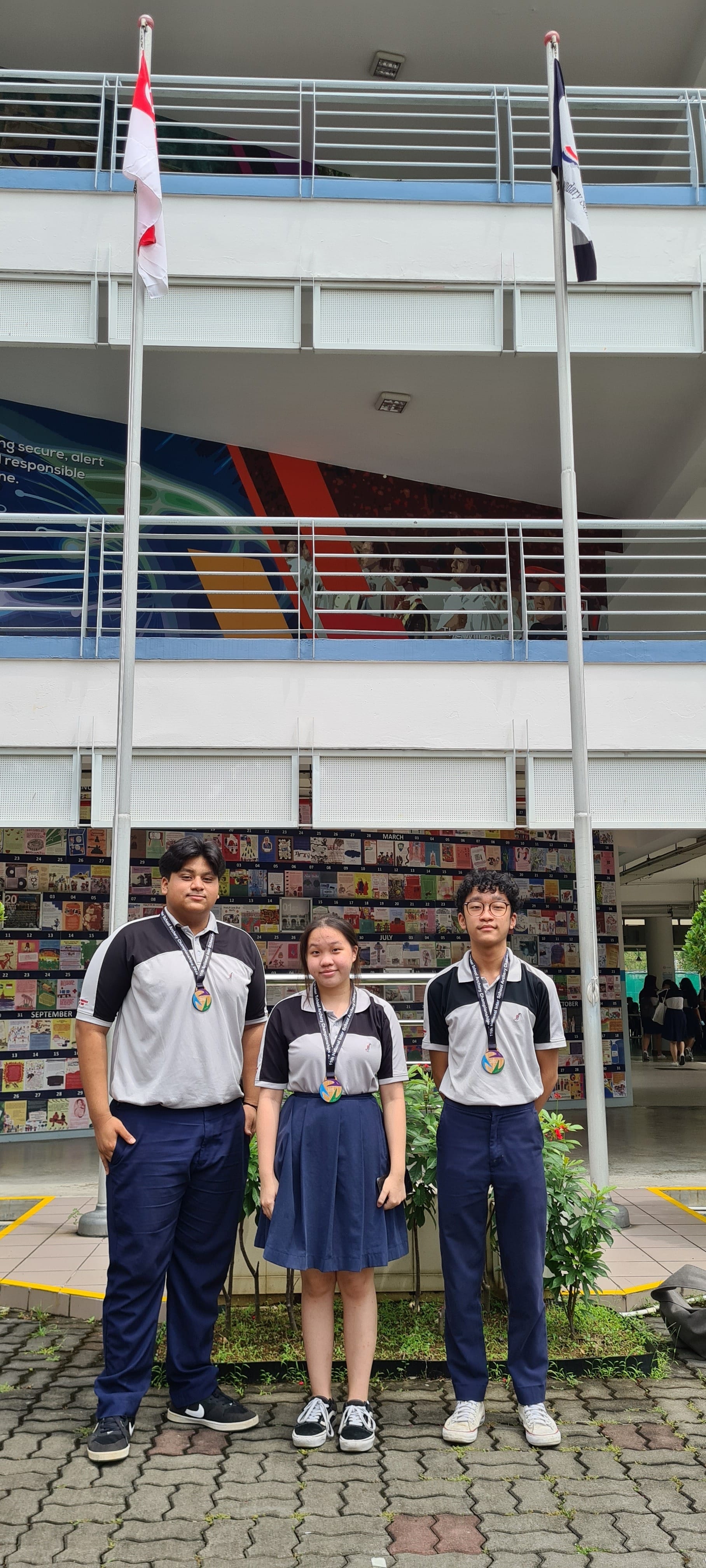 Three students in matching school uniforms and medals stand outdoors in front of a multi-story school building with flags.