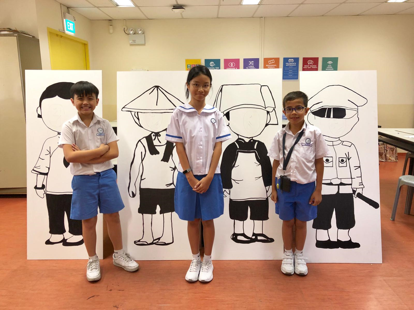 Three school children in uniforms stand in front of large boards with cartoon character cutouts in a classroom.