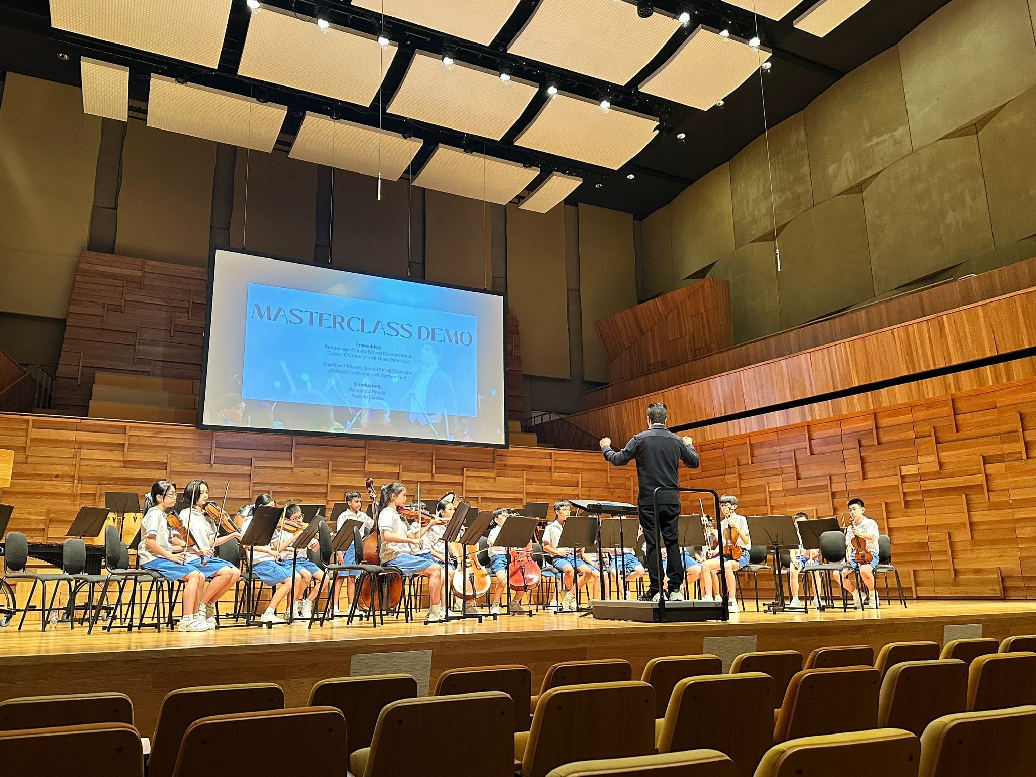 A conductor leads young student string players on a concert hall stage with a Masterclass Demo screen behind them.