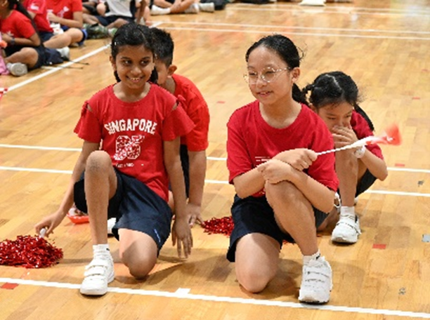 Children in red Singapore shirts kneel on a gymnasium floor, one holding a small flag and another with red pompoms.