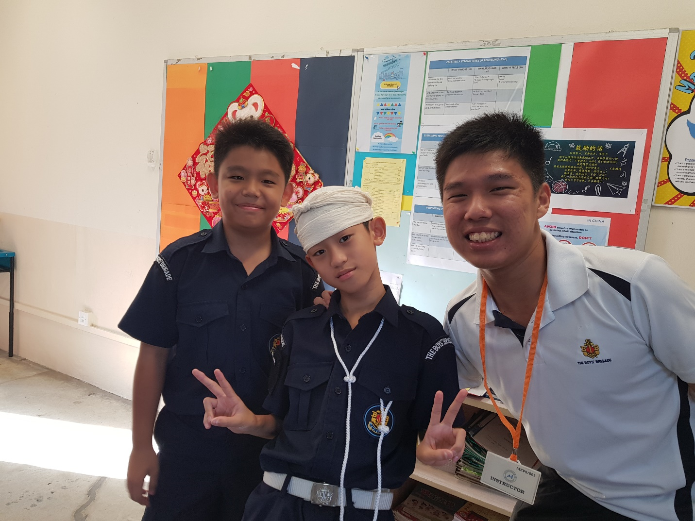 Two boys in navy uniforms and an adult instructor smiling and posing with peace signs in a classroom.