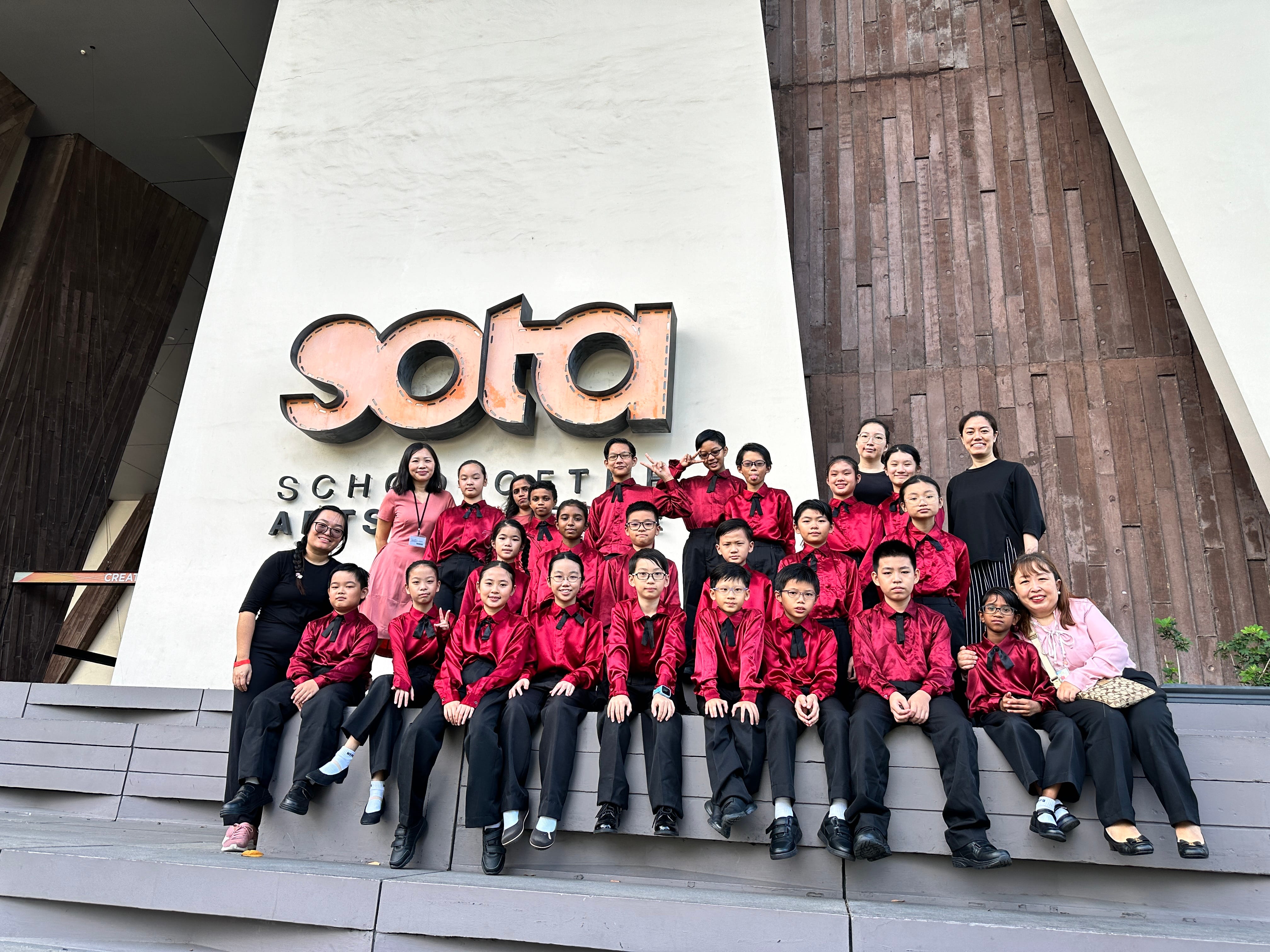 A group of children in red and black uniforms poses with adults in front of the SOTA School of the Arts building.