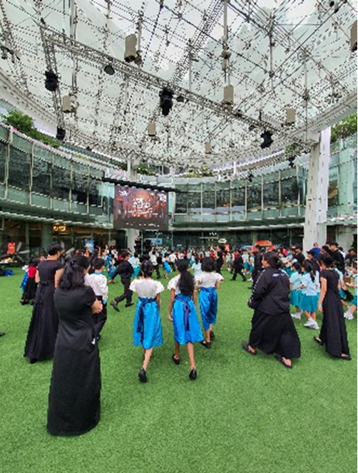 A large indoor atrium with a glass ceiling hosts a crowd of people on a green turf floor, some in blue skirts and black dresses.