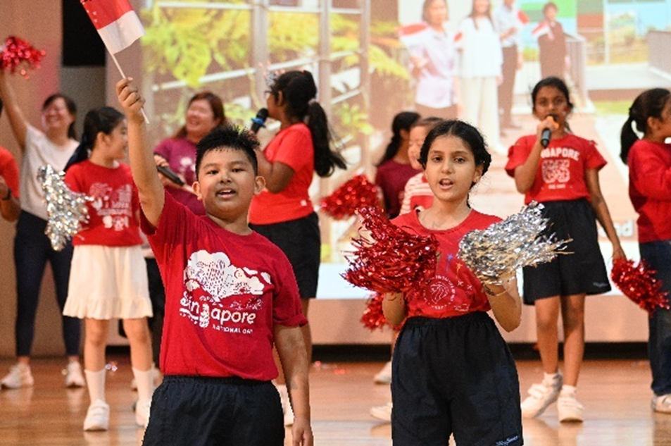 Children in red Singapore National Day shirts perform on stage, waving a flag and holding red and silver pom-poms.
