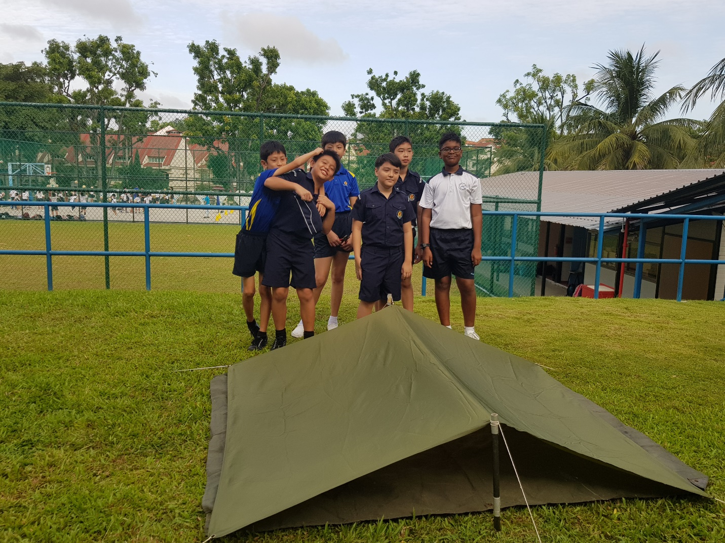 Five school students in uniforms stand on a grass field behind a green tent they assembled outdoors.