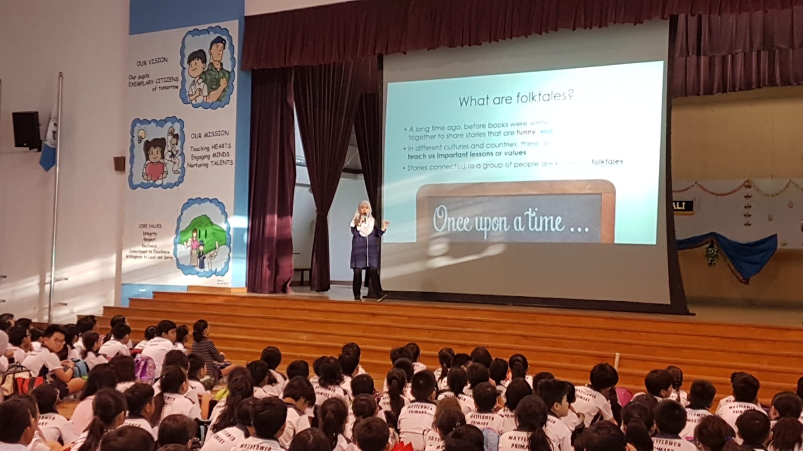 A speaker presents a folktales slideshow on stage to a large audience of uniformed primary school students in a hall.