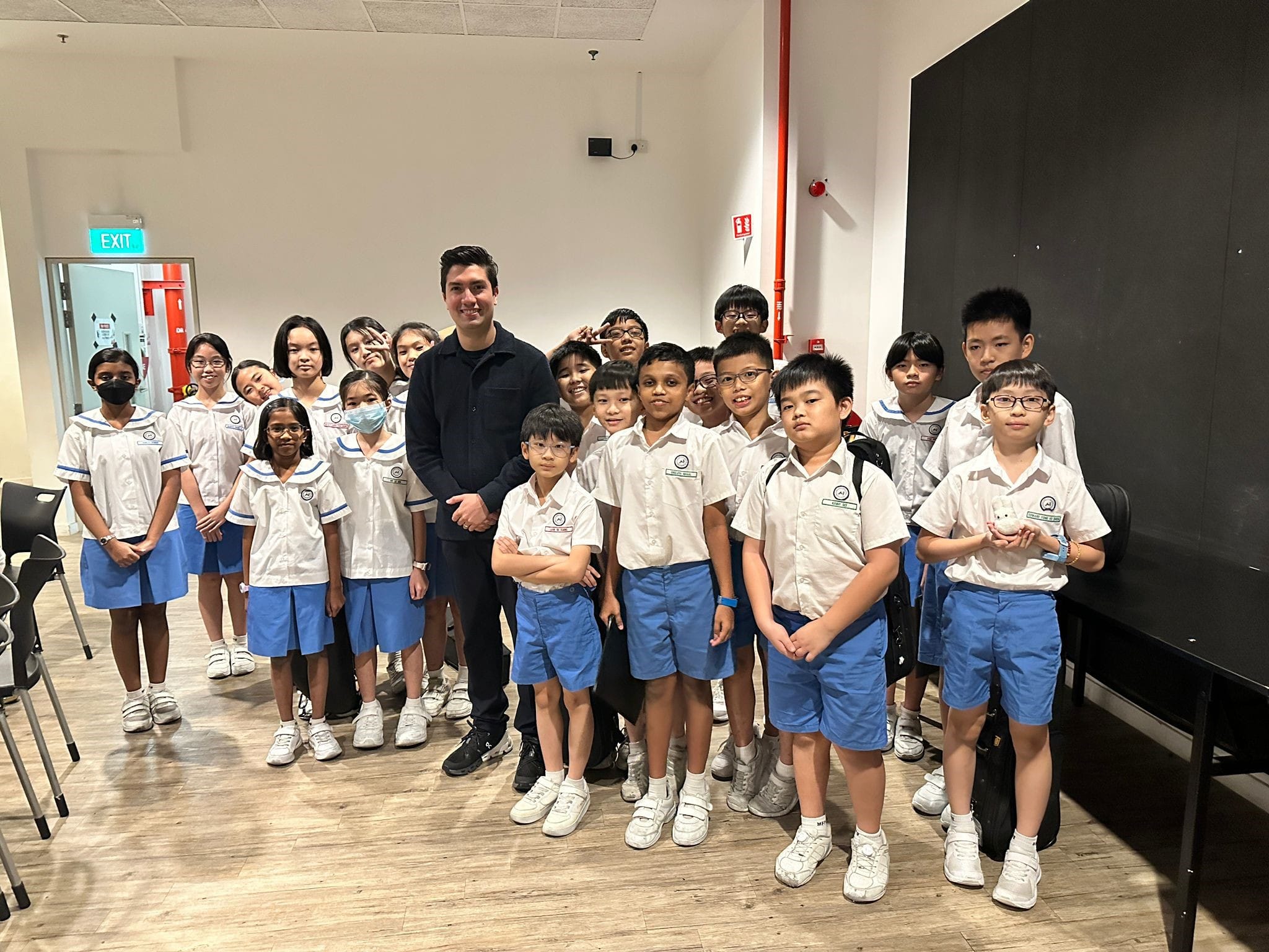 A adult man in black stands with a group of young students in white and blue school uniforms indoors.