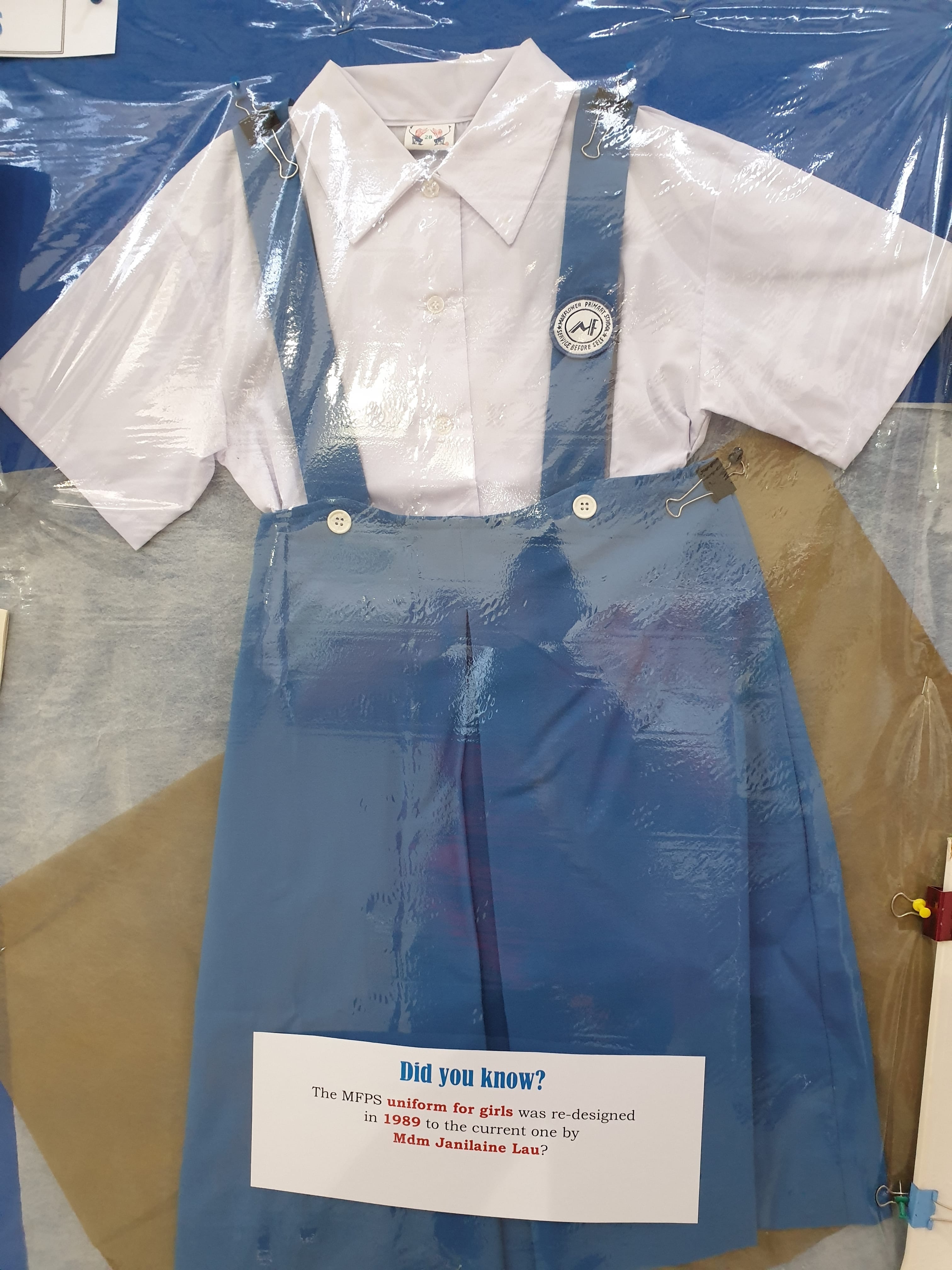 A displayed MFPS girls school uniform with a white shirt and blue pinafore, with a note about its 1989 redesign.