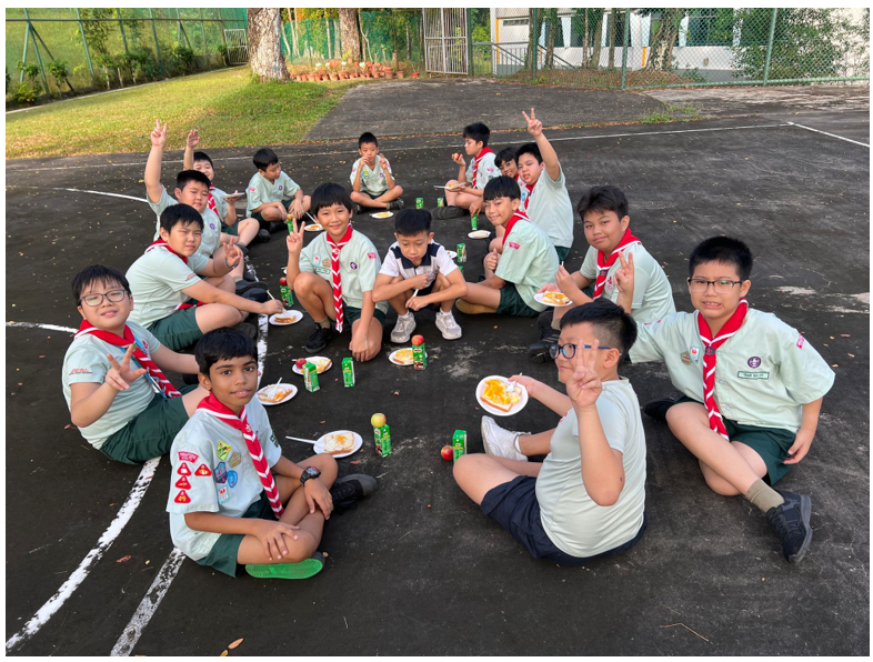 A group of young scouts in uniform sit on an outdoor court eating snacks and posing with peace signs.