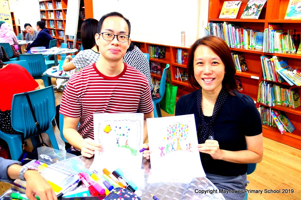 Two people smile while holding colorful drawings in a library filled with bookshelves and tables.