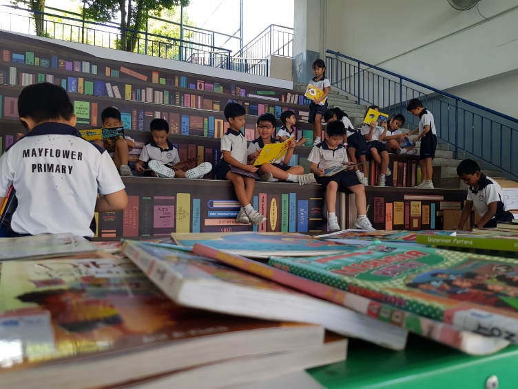 Mayflower Primary school children in uniform sit on steps reading books, with colorful books stacked in the foreground.