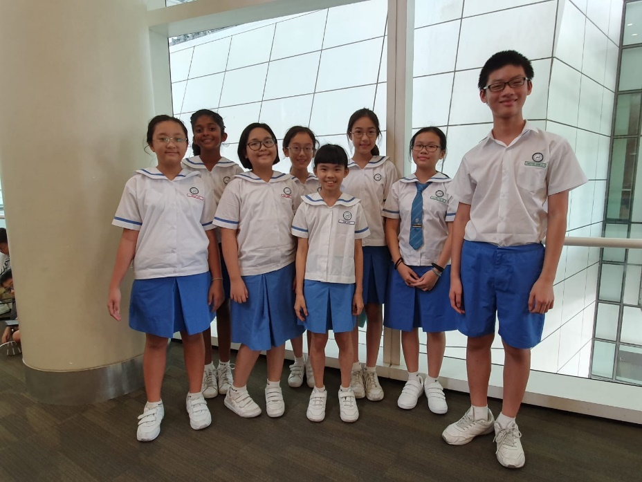 Eight students in white and blue school uniforms pose together indoors in front of large windows.