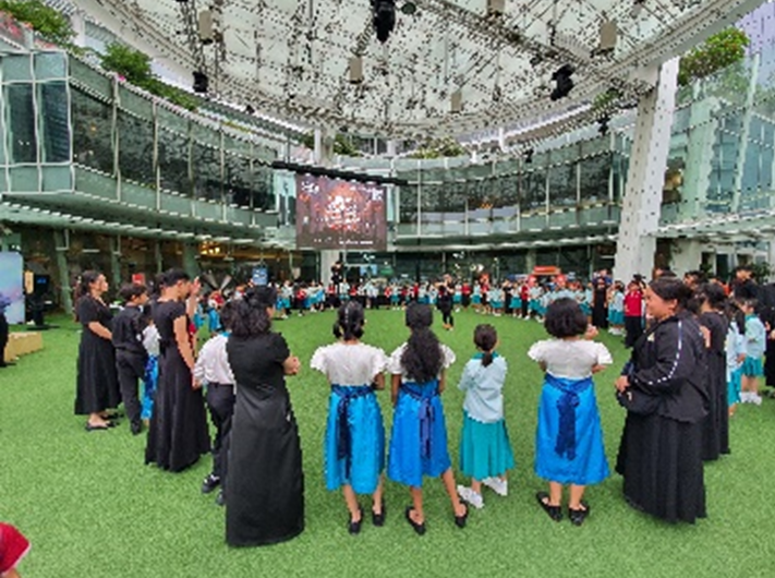 A group of performers in black and blue skirts stand in a circle on green turf inside a modern glass atrium.